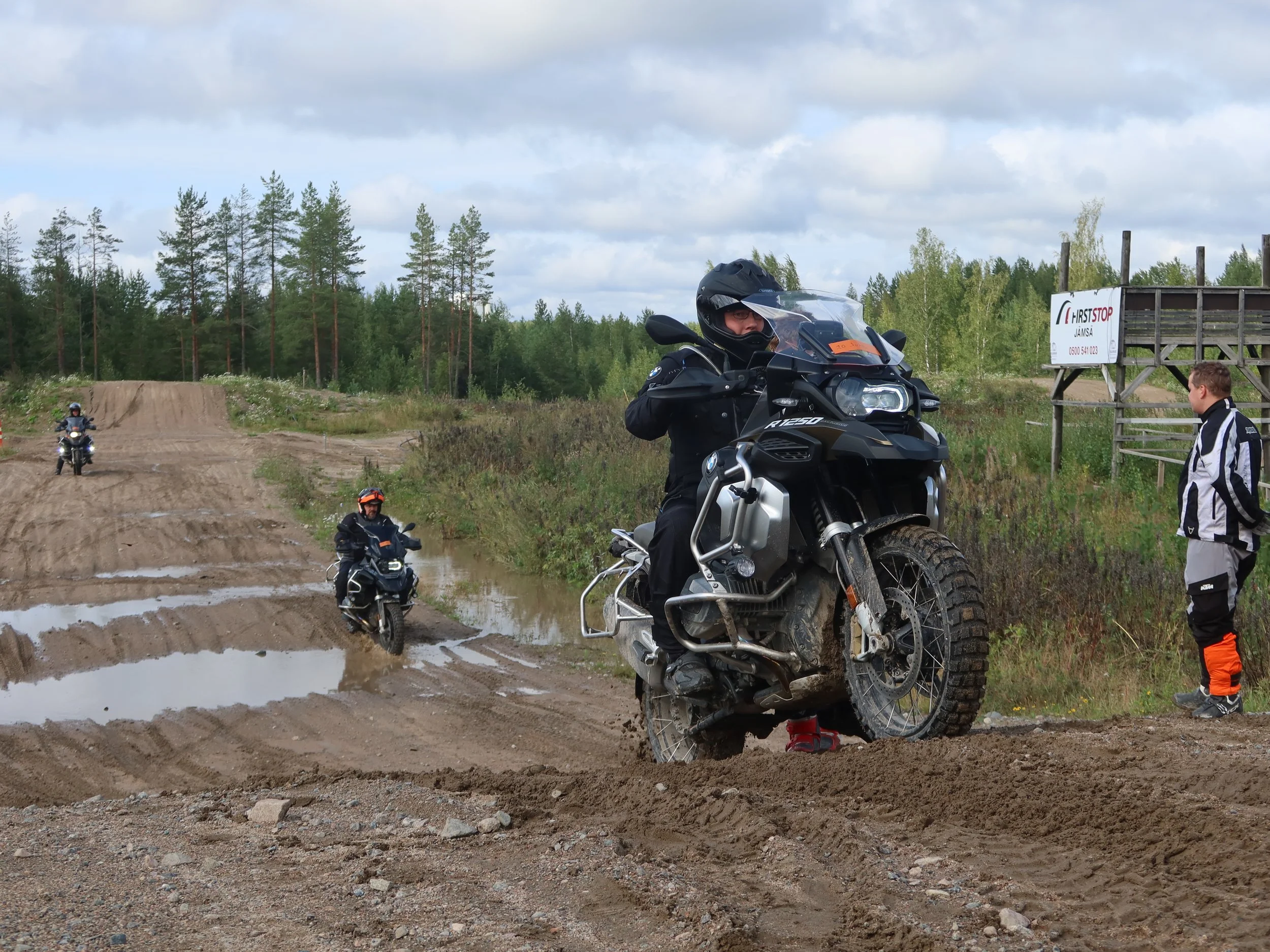 Miehellä on motocross-puku ja kypärä, hän istuu mustassa seikkailupyörässä. Taustalla kaksi muuta motoristia ja mies katselee oikealle. Maasto on mutainen ja märkä, maastopyörän renkaita on tahmeat. Taustalla on puista metsää ja pilvet taivaalla.