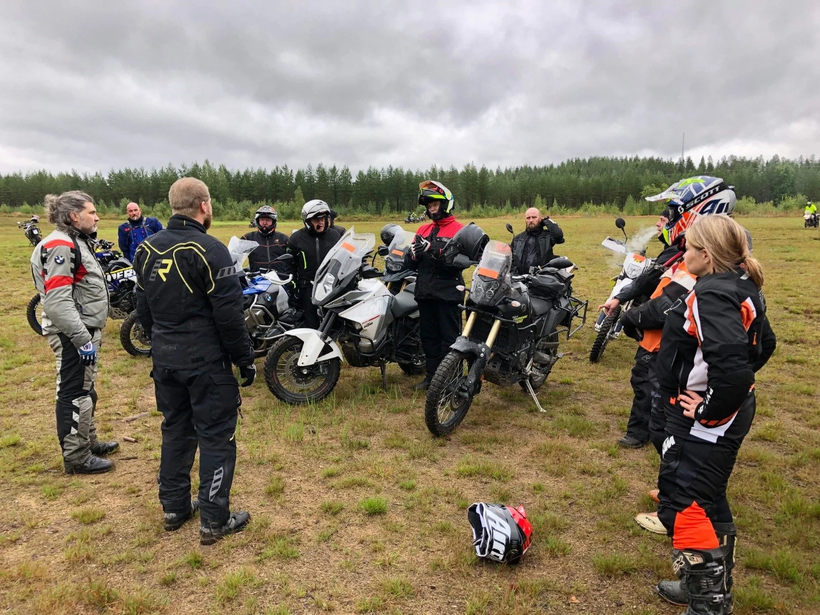 Ryhmä motocross-pyöräilykypärineitä ja -takkeja käyttäviä ihmisiä keskustelemassa avoimella kentällä, pilvinen sää, vuoristomaisema