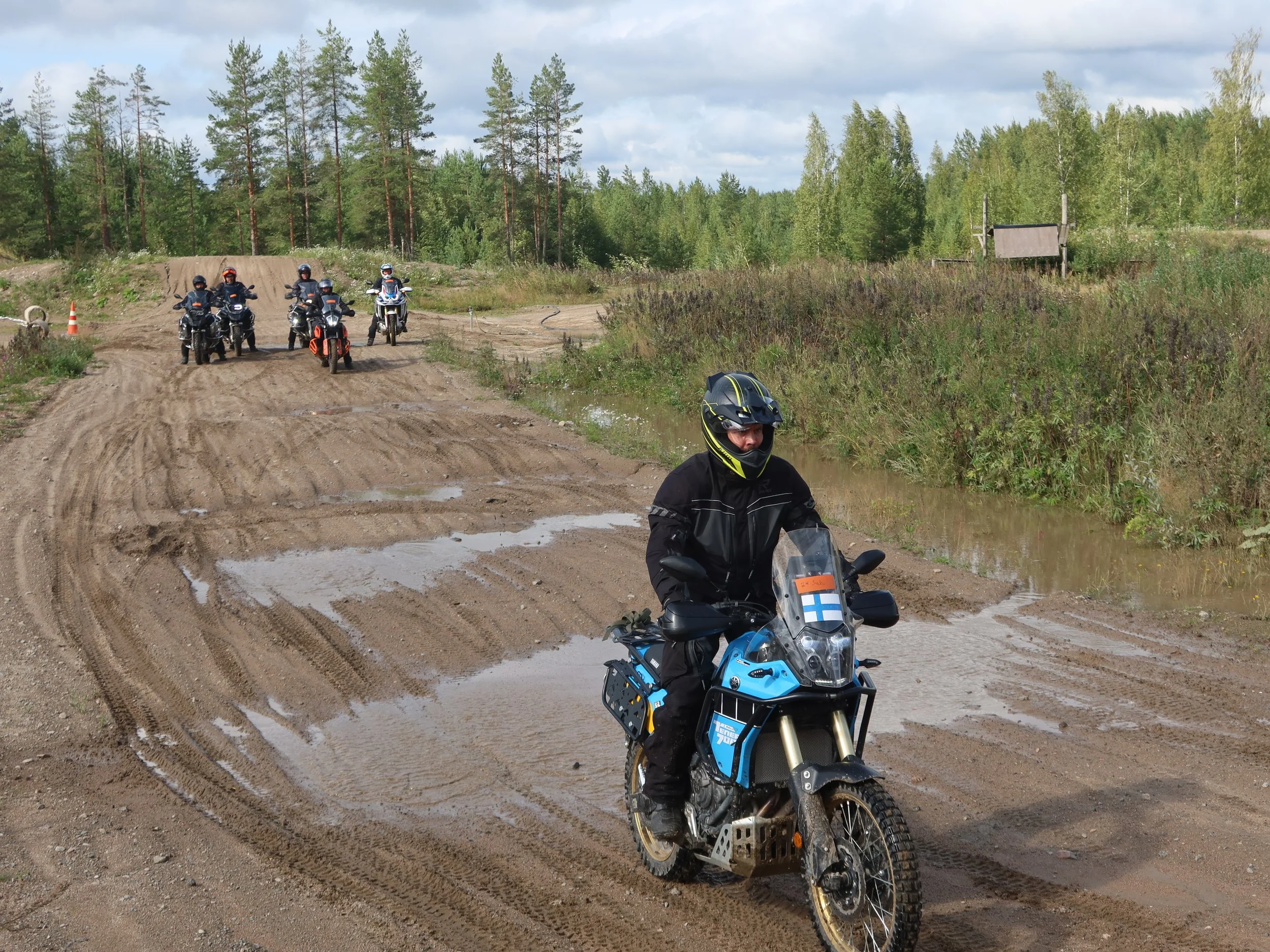 Miehet moottoripyöräilyssä metsäisessä maastossa, yksi etualalla ja useita takanapäin, maasto on mutainen ja märkä, ympärillä puita ja luonnonäkymää.