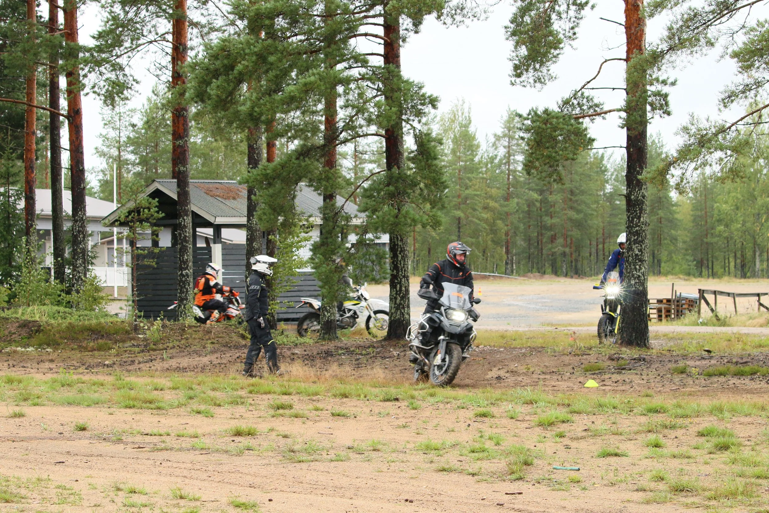 Neljä moottoripyöräilijää harjoittelee maastossa, ympärillä puita ja rakennuksia