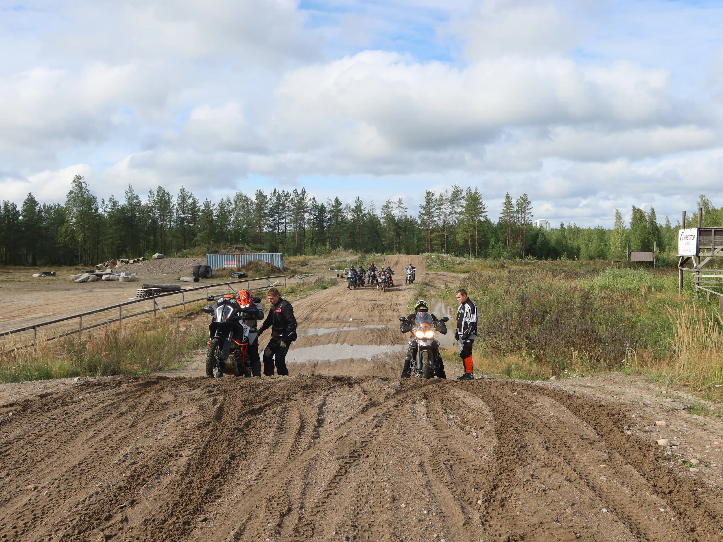 Motoristit ja mopot pysähtyneinä hiekkatien varrella, liikennemerkki oikealla, taustalla metsä ja pilvinen taivas.