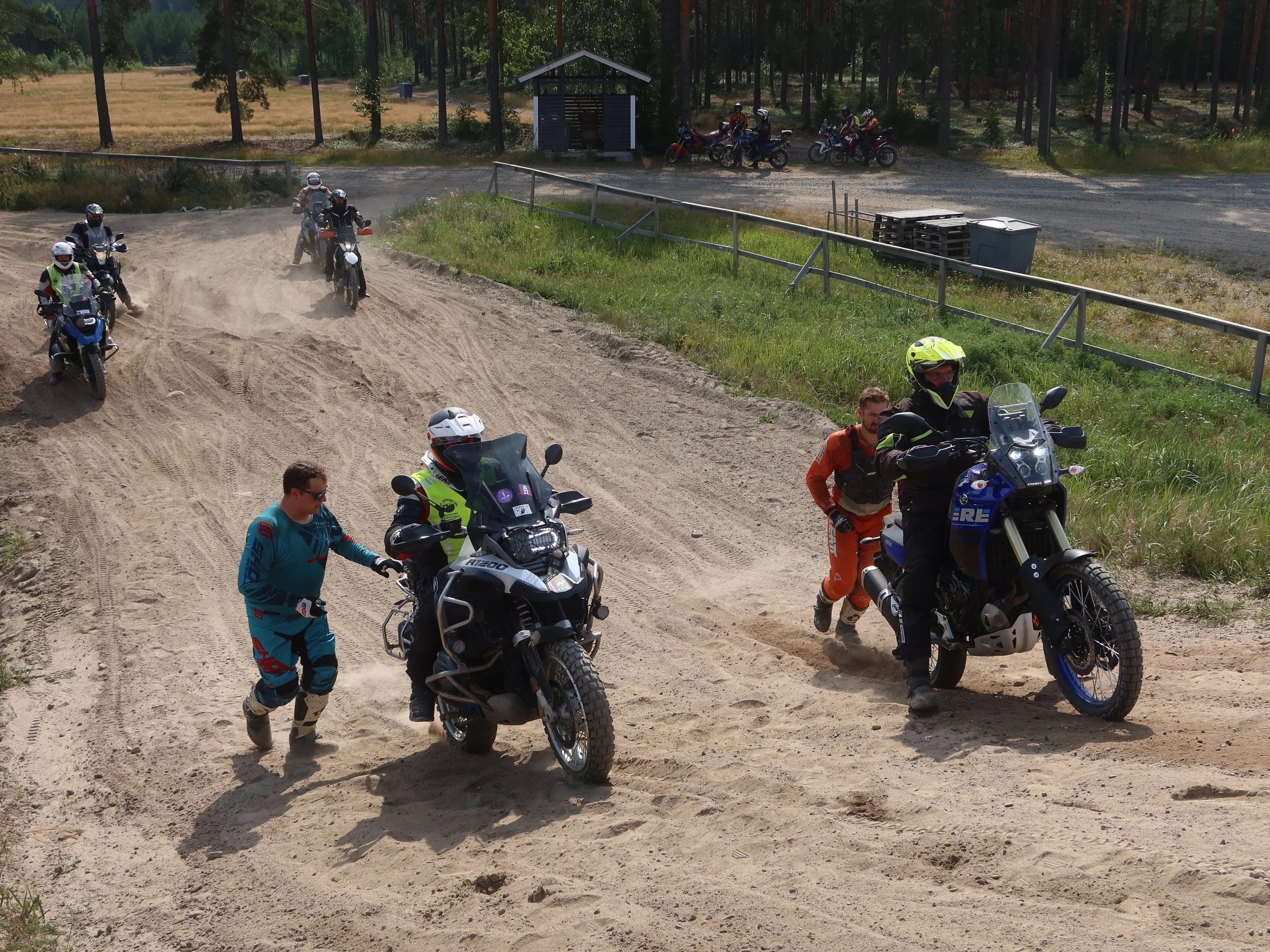 Henkilöitä siirtämässä motocross-konetta ylös mäkeä, muita motoristeja odottamassa taustalla, hiekkatie ympäristö.