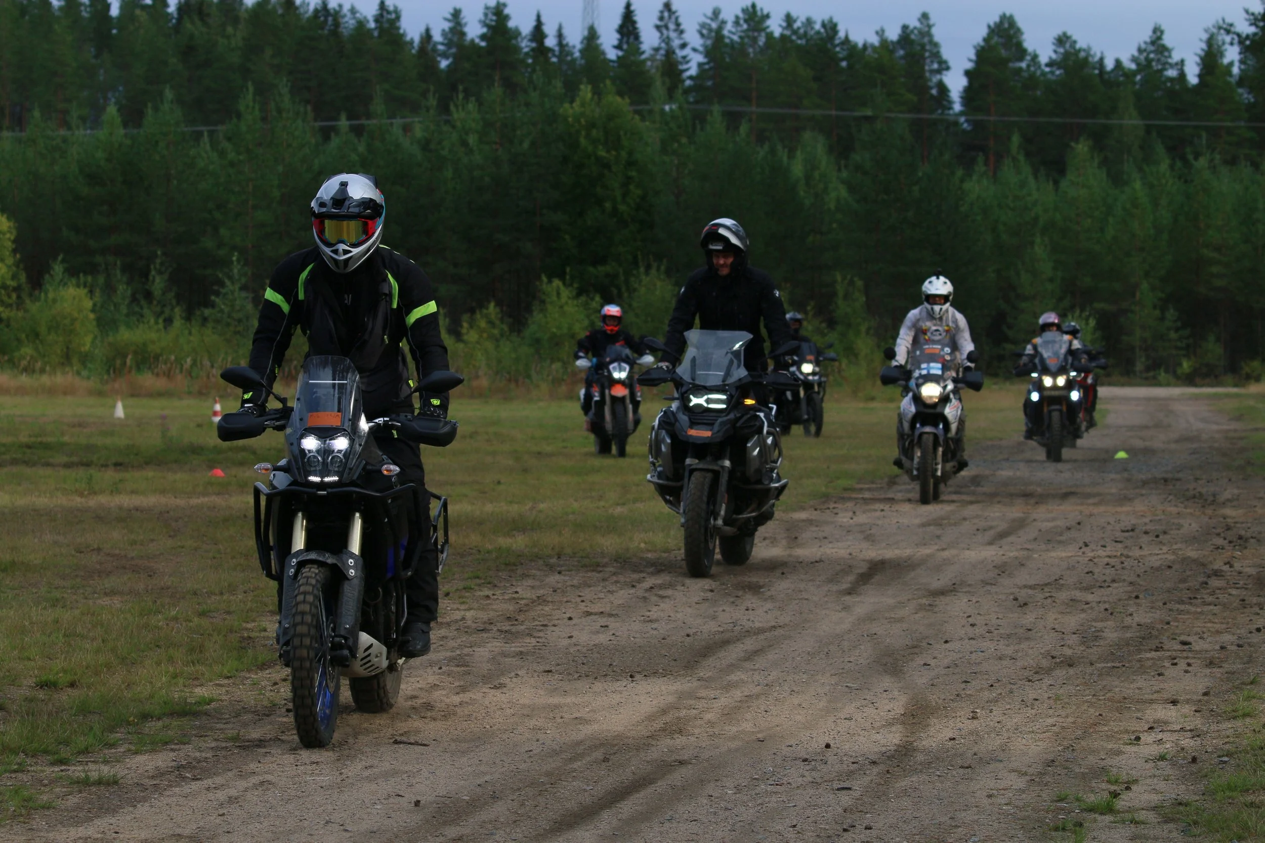 Ryhmä motoristeja ajamassa maastossa, ensimmäisenä valkoiset kypärät ja mustat ajovarusteet, taustalla vihreitä puita.