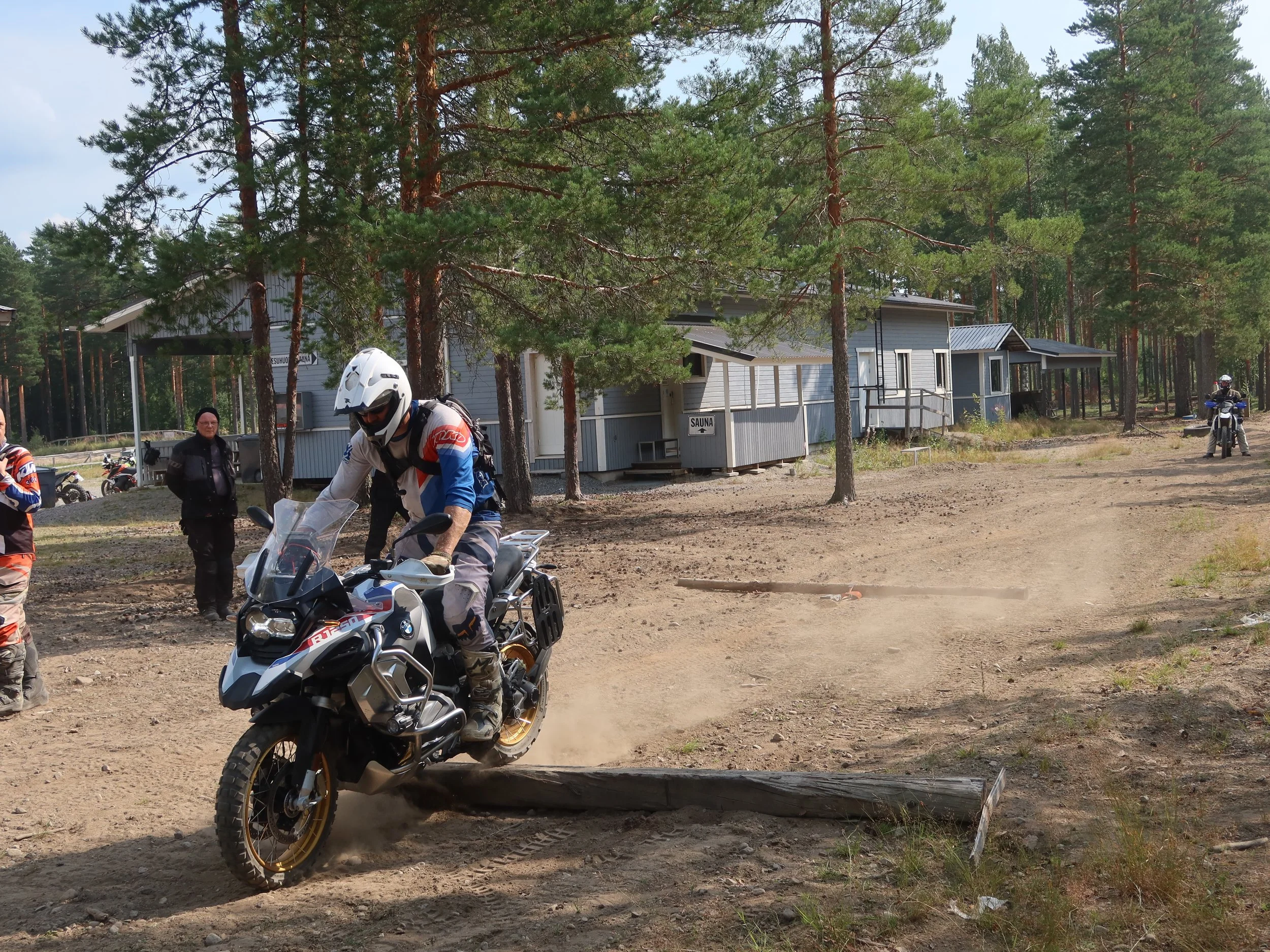 Mies motoripyörällä esteen yli maastossa, taustalla muita moottoripyöräilijöitä ja rakennuksia puitten ympäröimänä.