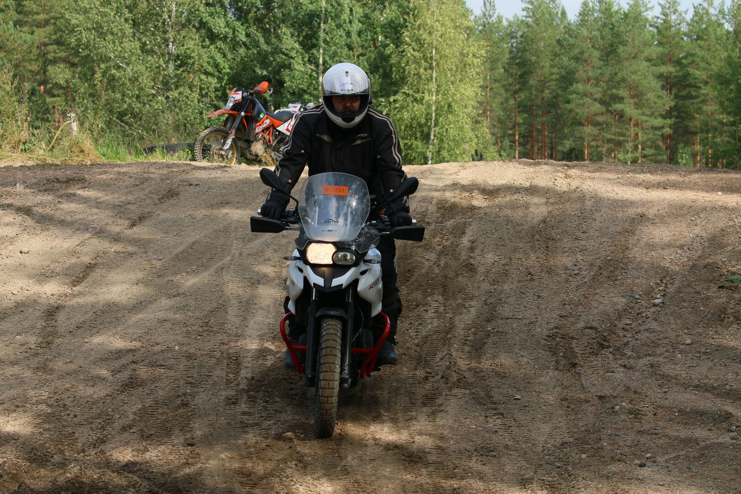 Mies ajoa motocross-pyörällä metsäisessä maastossa, toinen motocross-pyörä ja ihmisiä taustalla.