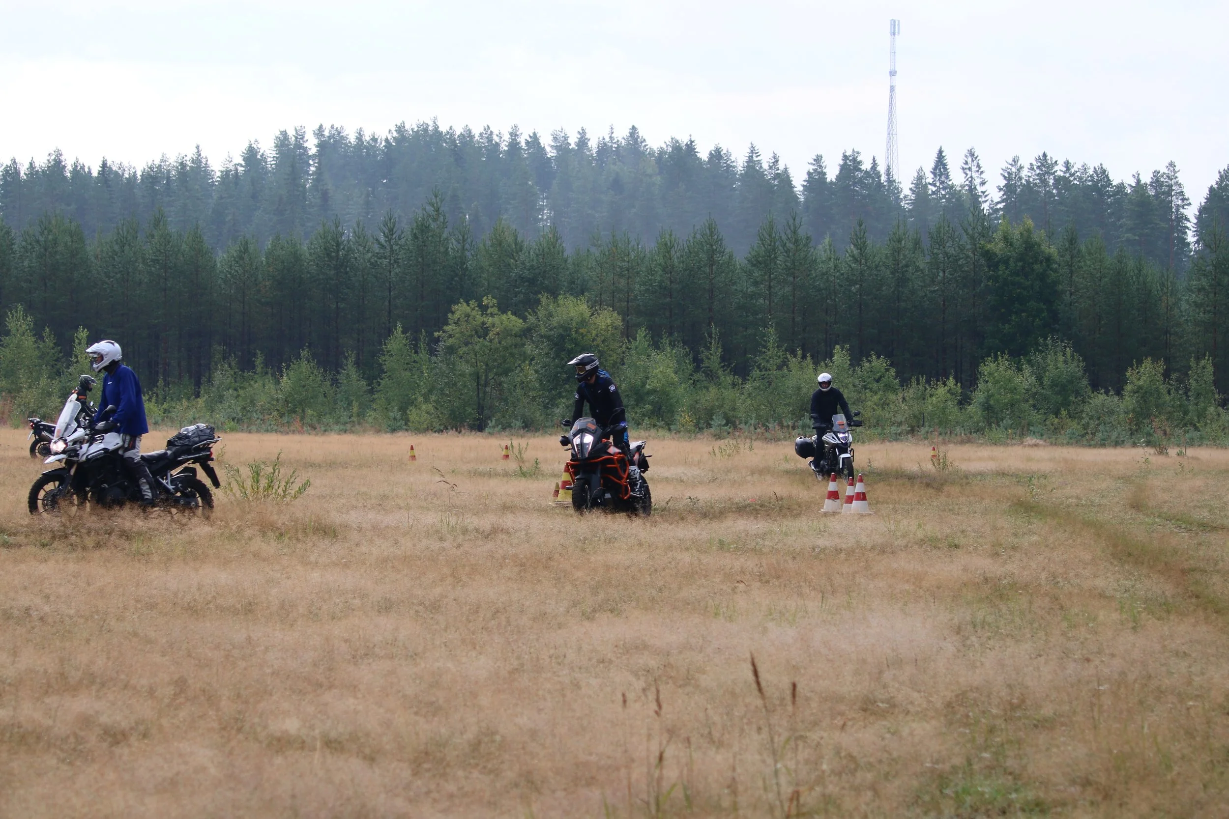 Kolme moottoripyöräilijää lähellä vihreitä puita ja sinistä taivasta, harjoitellen ajoneuvotaitojaan hiekkakentällä, joissa on liikennemerkkimäisiä merkkejä ja roiskuvaa hiekkaa.