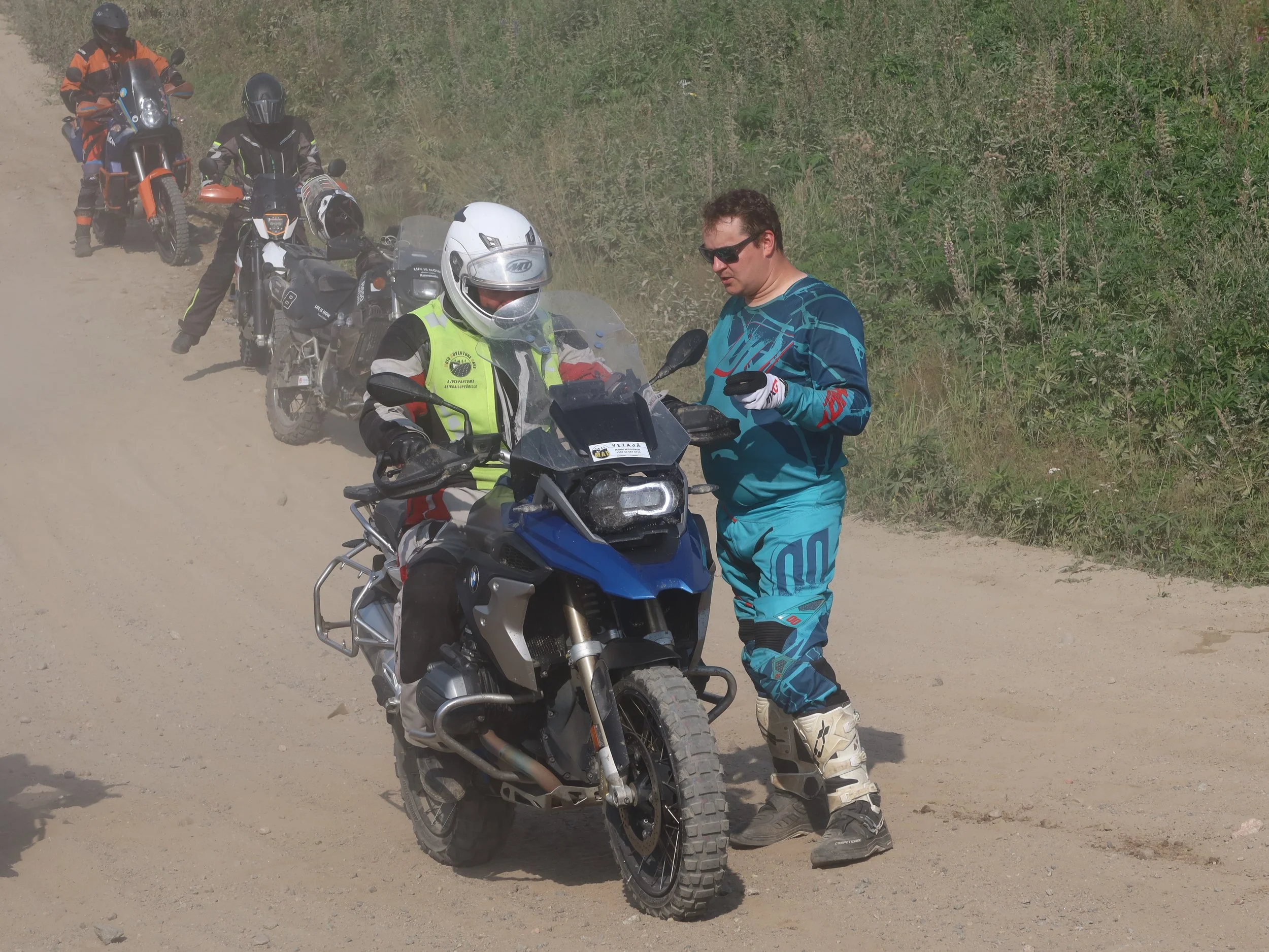 Ryhmän motocross-kuskit pysähtyneet hiekkatievaelluksen aikana keskustelemaan, mies puvussa ja kypärässä istuu moottoripyörällä, toinen mies seisoo rinnalla näyttäen puhelintaan.
