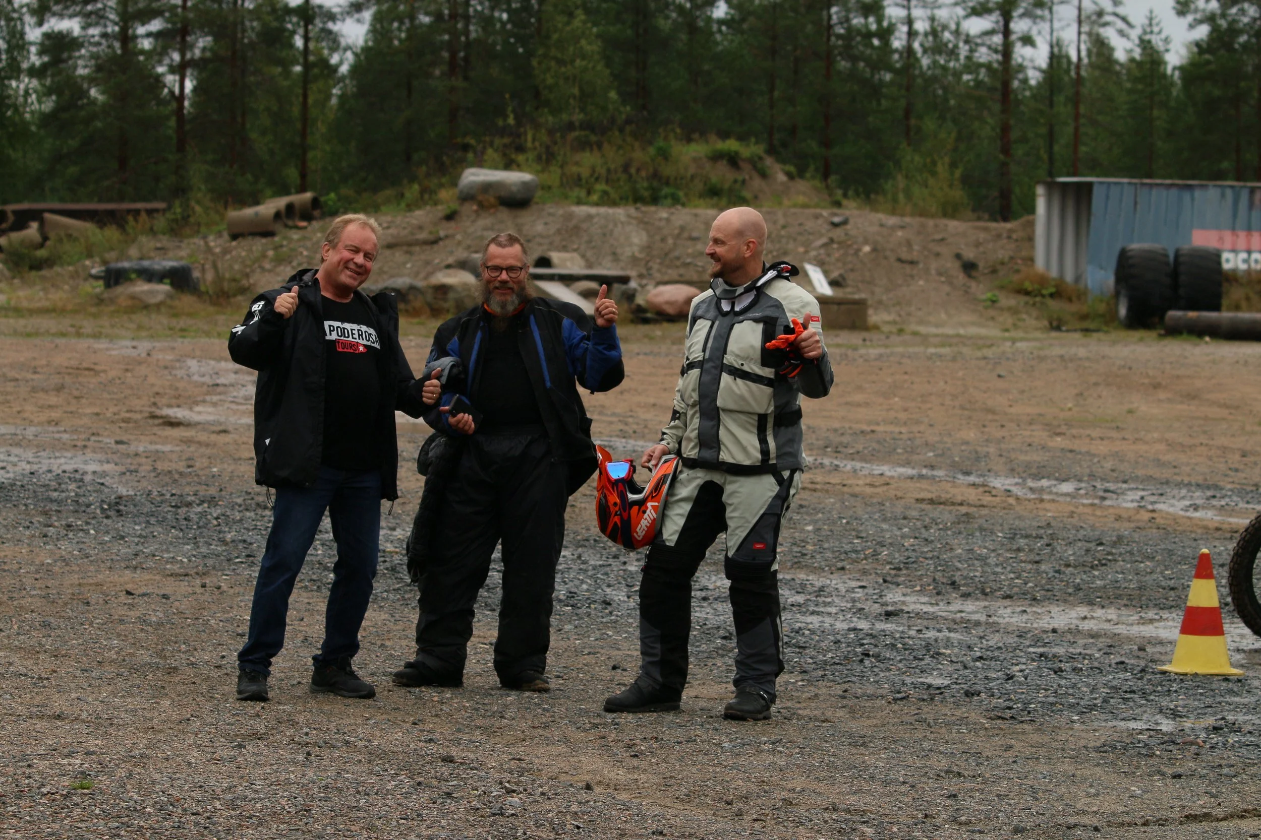 Kolme miestä, jotka on pukeutunut motocross- ja offroad-varusteisiin, seisovat ja juttelevat ulkona, taustalla on metsää, kuoppaisia teitä ja renkaita.