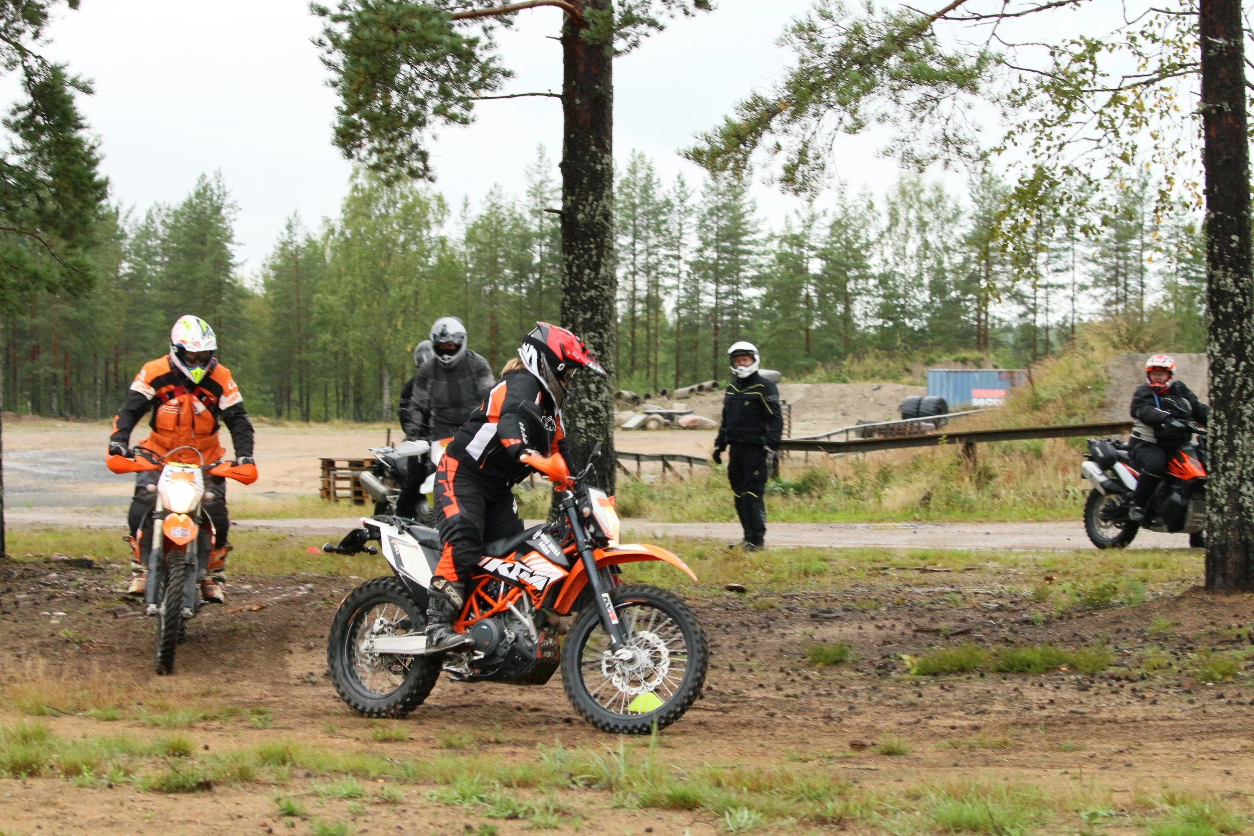 Neljä motocross-harrastajaa ja kaksi valvojaa, kaikki kypärissä ja suojavarusteissa, harjoittelemassa metsäisellä radalla