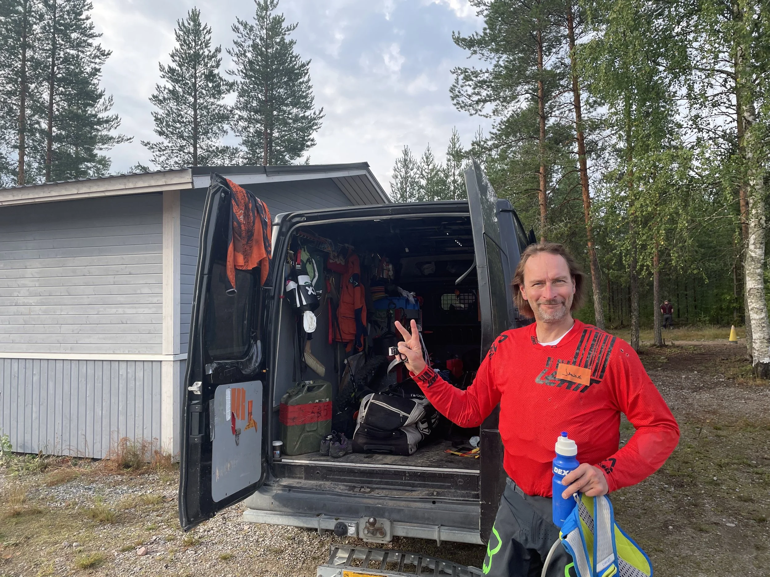 Mies hymyilee ja tekee rauhallisen ilmeen, seisoo ulkona pyöräilyvarusteiden kanssa, jonka vieressä on avoin pakettiauto täynnä varusteita, taustalla metsä ja pieni rakennus.