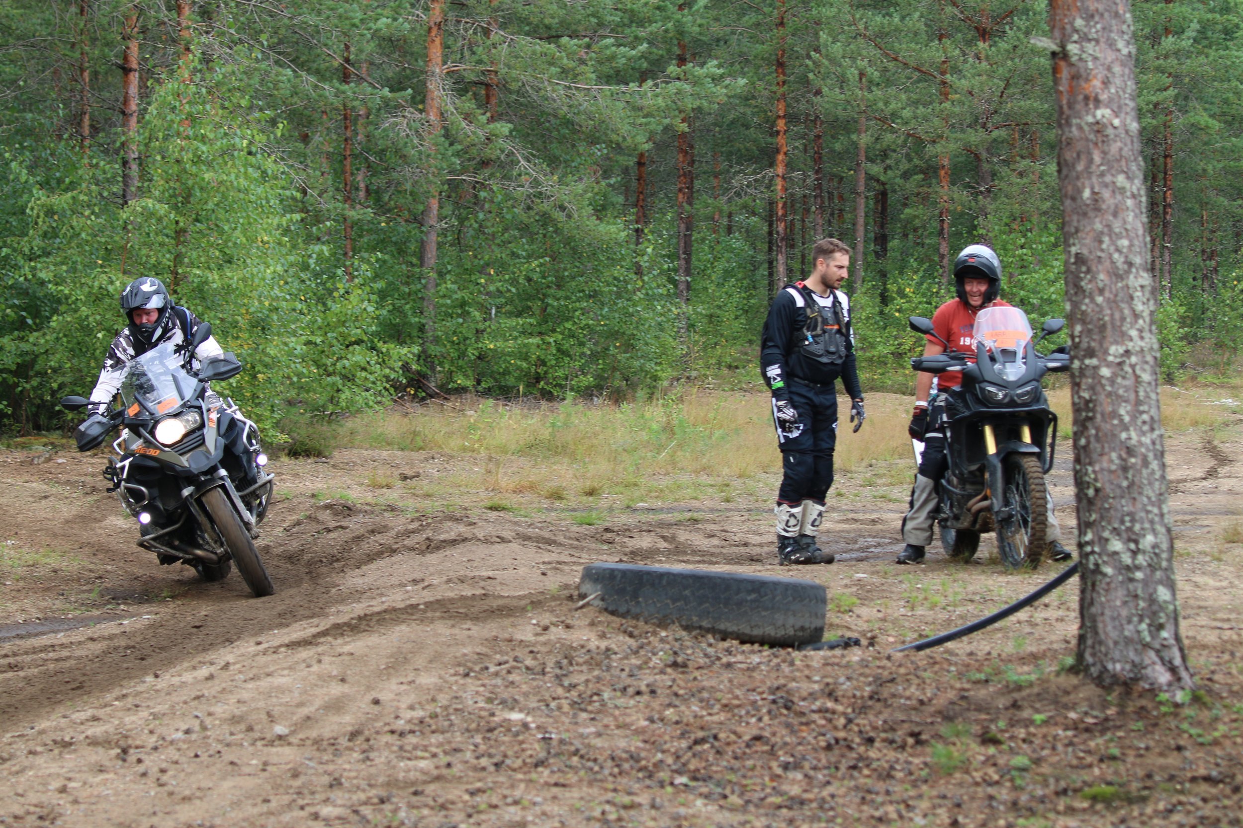 Kaksi motocross-harrastajaa ja yksi motocross-mopot, yksi istuu maassa ja kaksi seisoo metsässä, taustalla puita ja nurmikkoa.