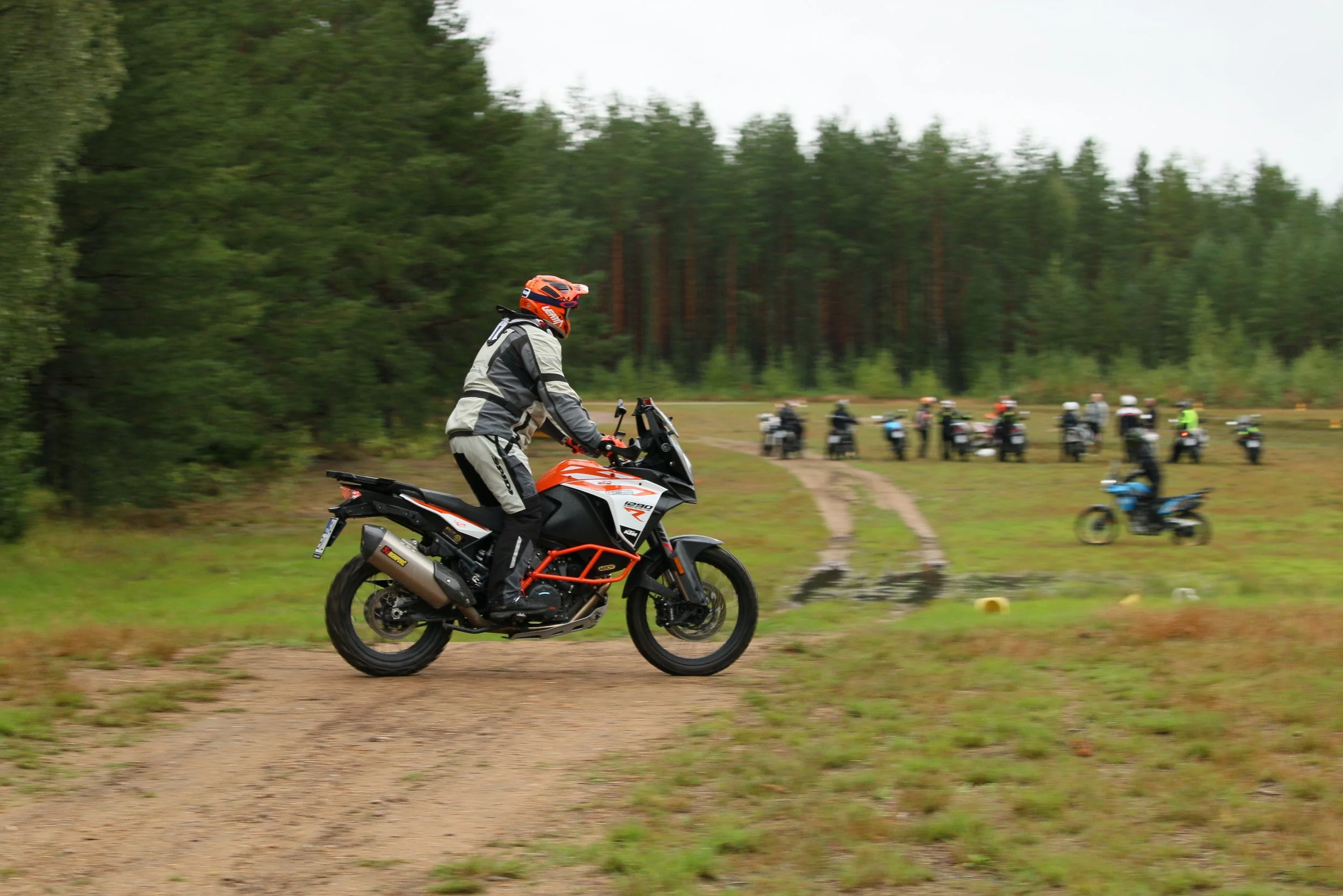 Mies ajamassa maastopyörällä metsäalueella, taustalla muita motoristeja ja polkupyörä, ympäröivä metsä ja mutainen polku.