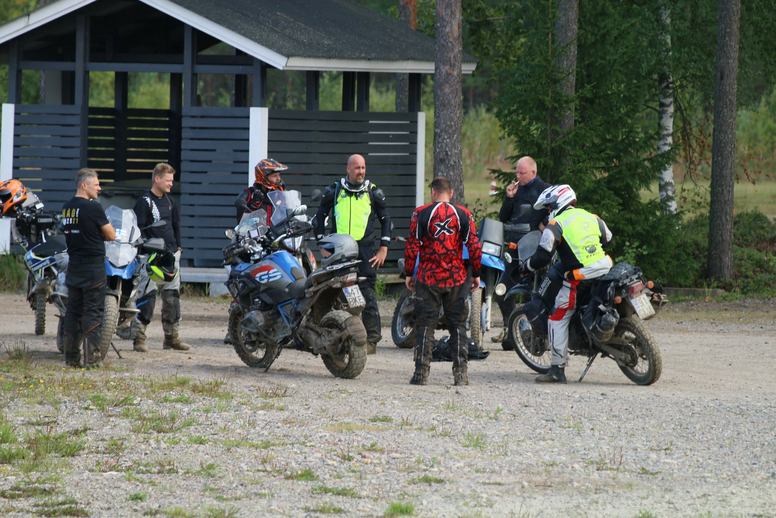 Seitsemän motocross-kypärä ja maastopyöräkerhoa ajavaa henkilöä kokoontuu keskusteluun metsän vieressä ja parkkipaikalla