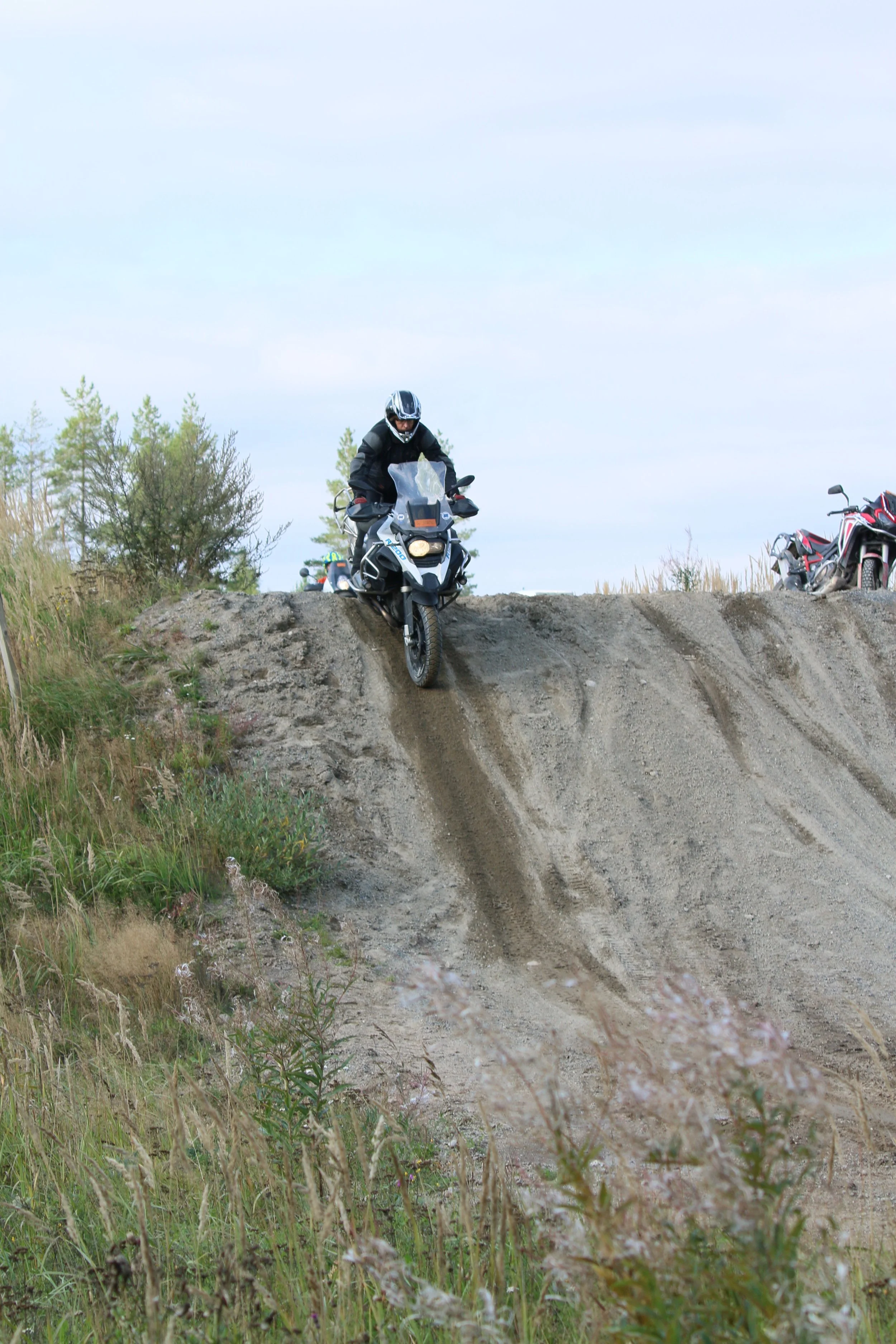 Mies ajaa motocrossilla ylämäkeä ramppia pitkin kyläilössä ja muita motoristeja odottelee yläpuolella.