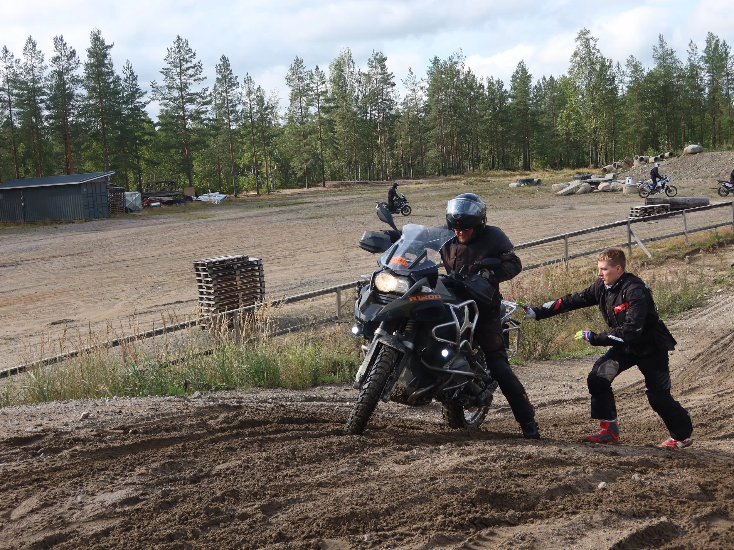 Mies ajoje motocrosspyörällä ja toinen henkilö auttaa tai ohjaa hänet, külkipaikka ja metsää taustalla.
