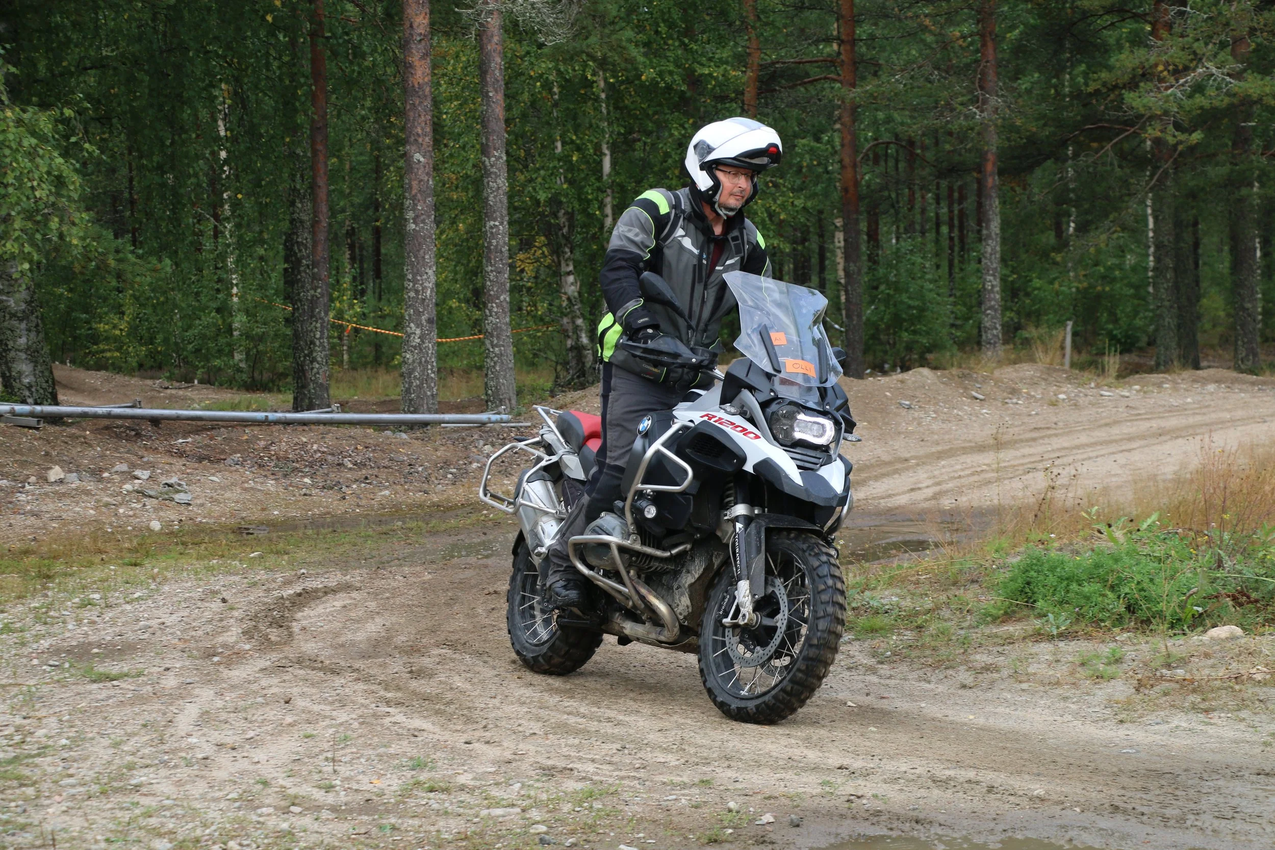 Mies kuljette maastoajo-polkua BMW R1200GS -moottoripyörällä metsän keskellä, ympärillä puita ja hiekkatie.