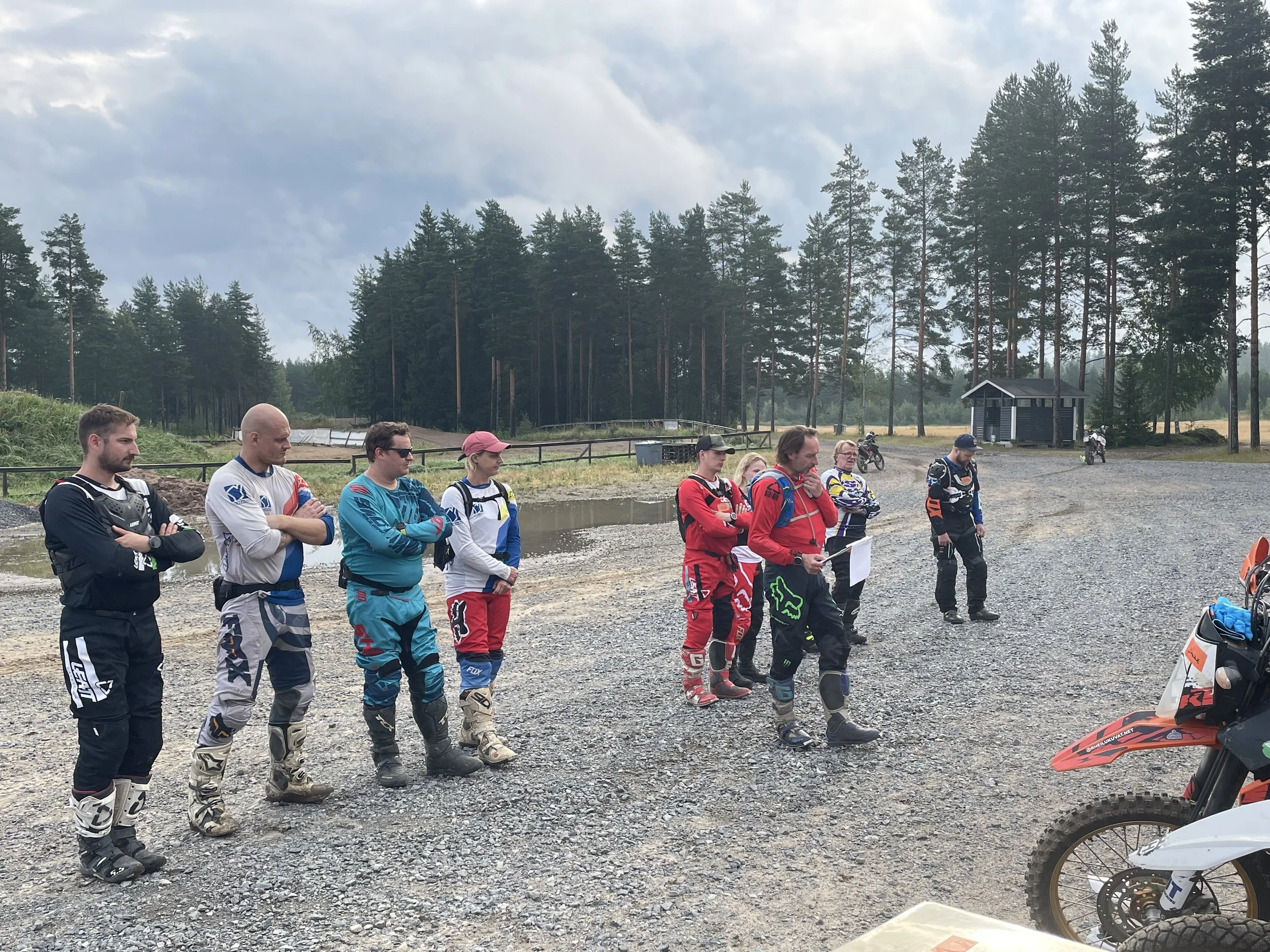 Motorcross-kilpailuun osallistuva joukko ihmisiä odottaa yhteislähdössä rinteessä, taustalla metsää ja pieni rakennus.