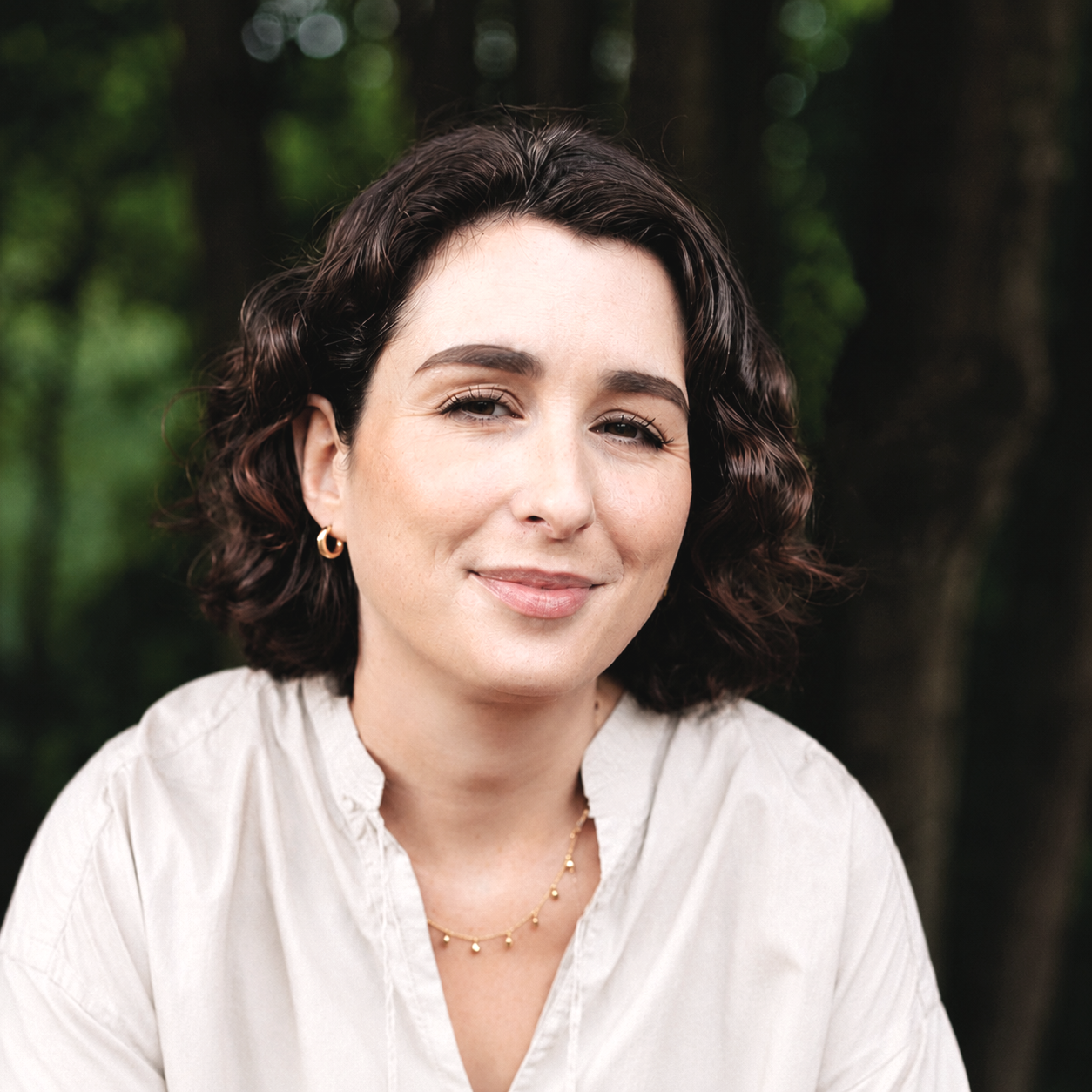 Portrait of a woman with short, curly dark hair, wearing gold hoop earrings, a delicate gold necklace, and a light-colored blouse, smiling softly with a blurred green forest background.