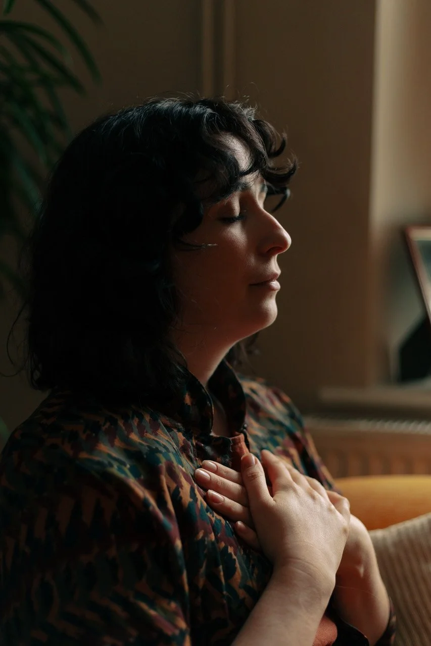 A woman with dark, wavy hair and closed eyes is resting her hand on her chest, in a warm indoor setting near a piano.