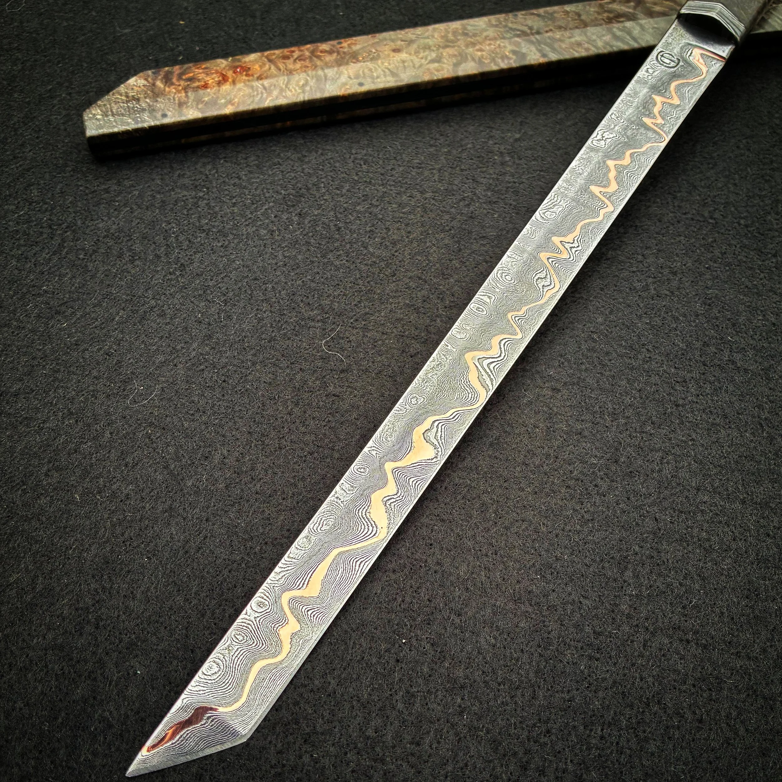 Close-up of a Damascus steel kitchen knife with a decorative patterned blade and a textured wooden handle, resting on a black surface.