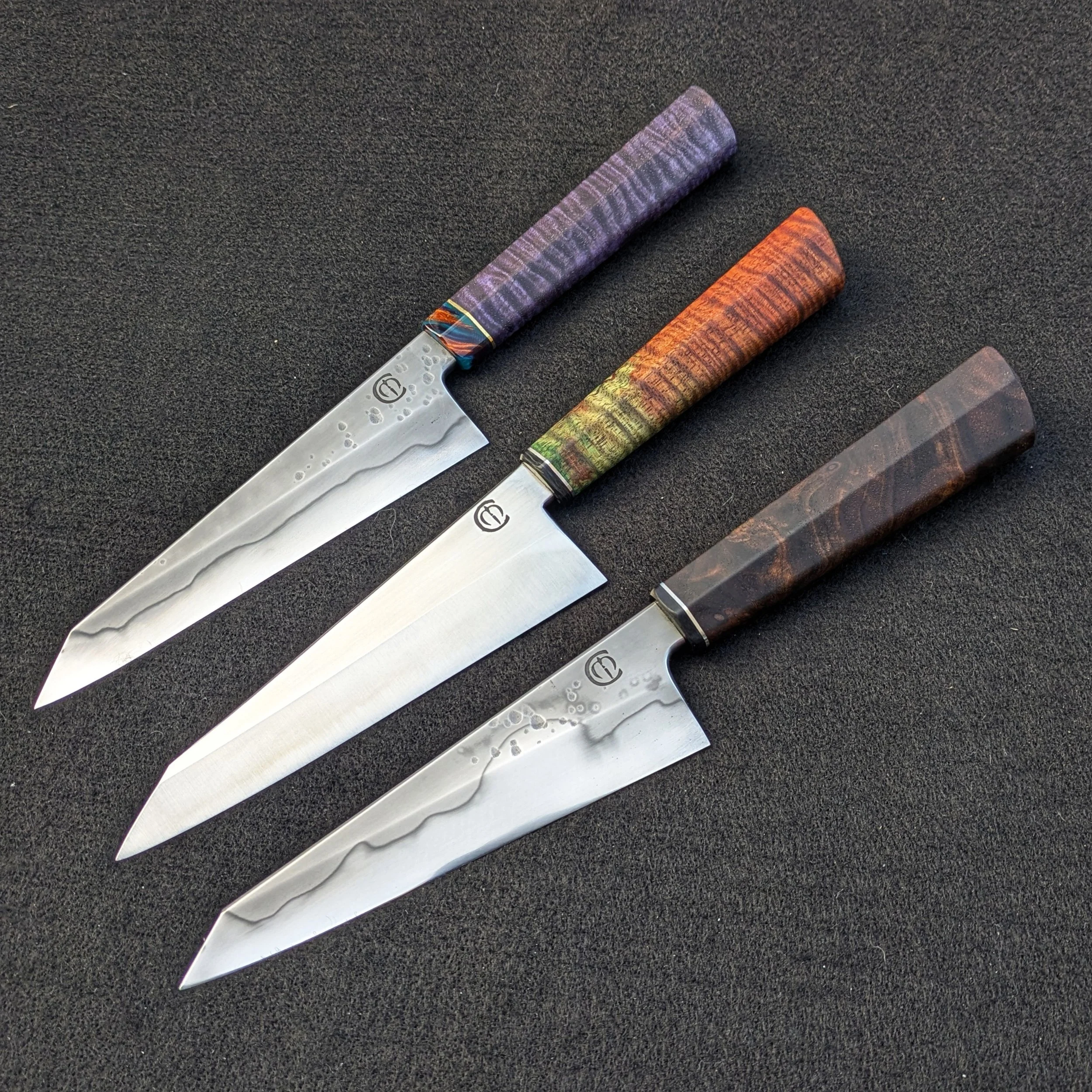 Three handcrafted kitchen knives with Damascus steel blades and colorful, patterned handles placed on a black surface.