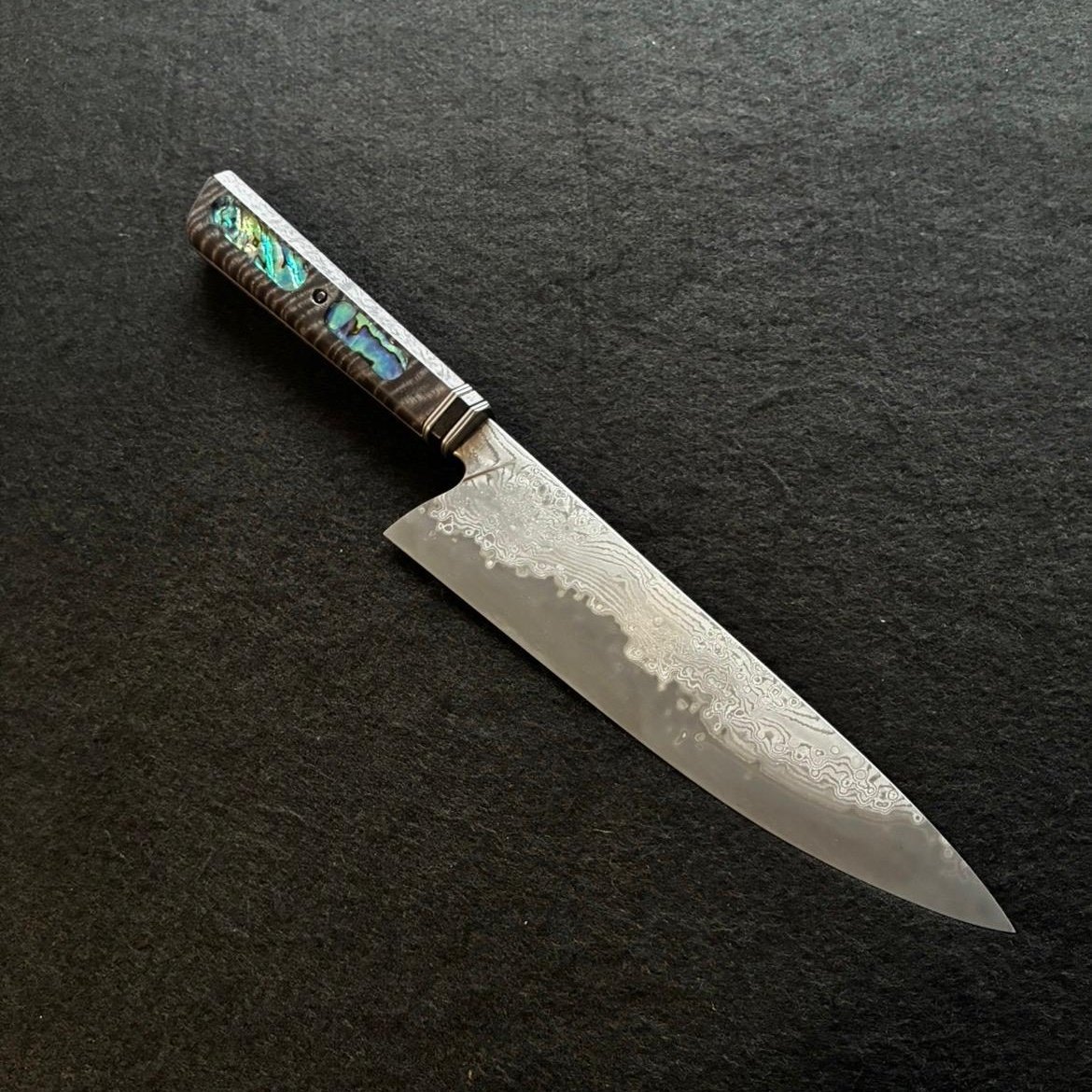 A decorative kitchen knife with a inlaid Abalone shell handle and a VG10 Suminagashi steel blade on a black surface.