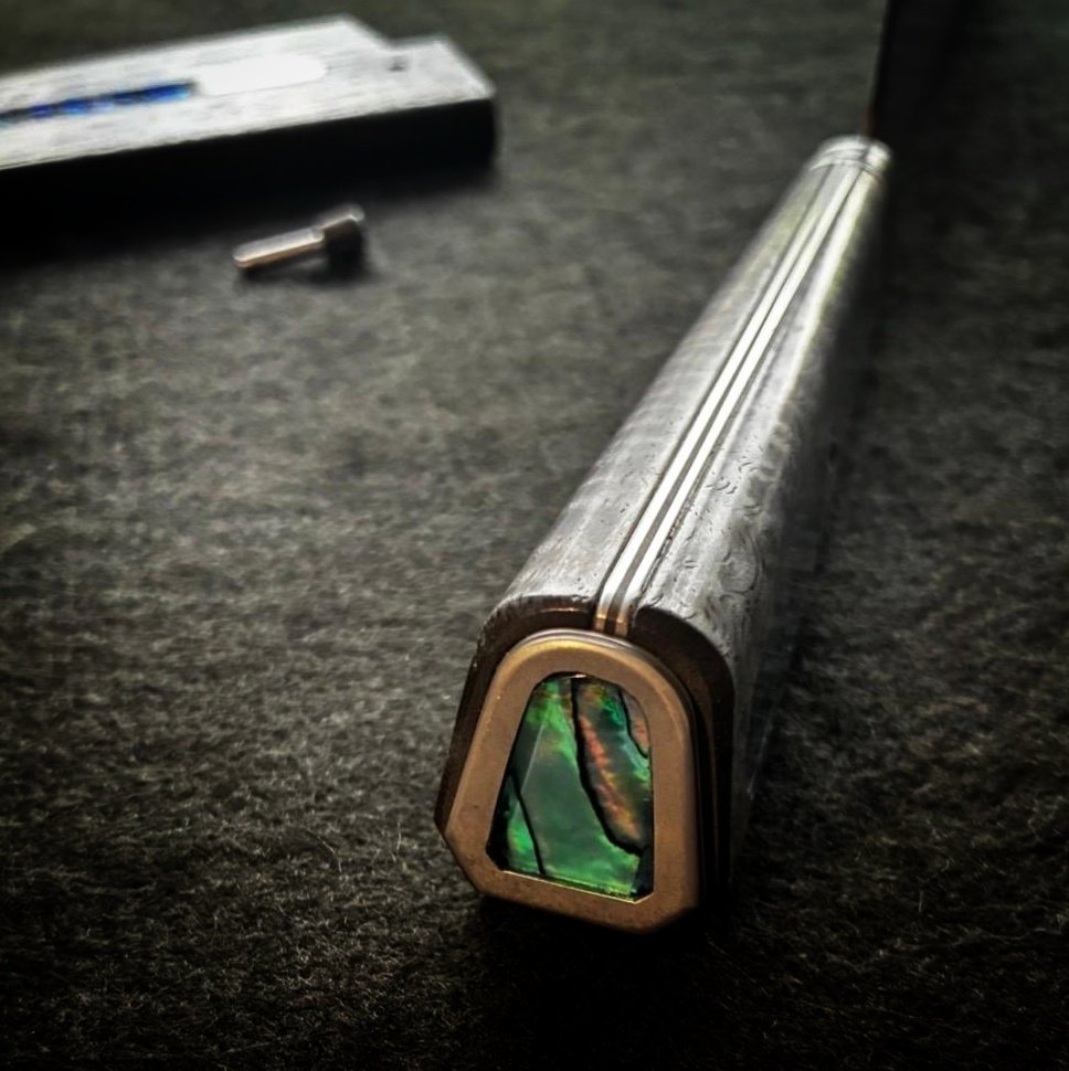 A close-up of a knife handle with an inlaid green and black abalone shell, lying on a dark surface.
