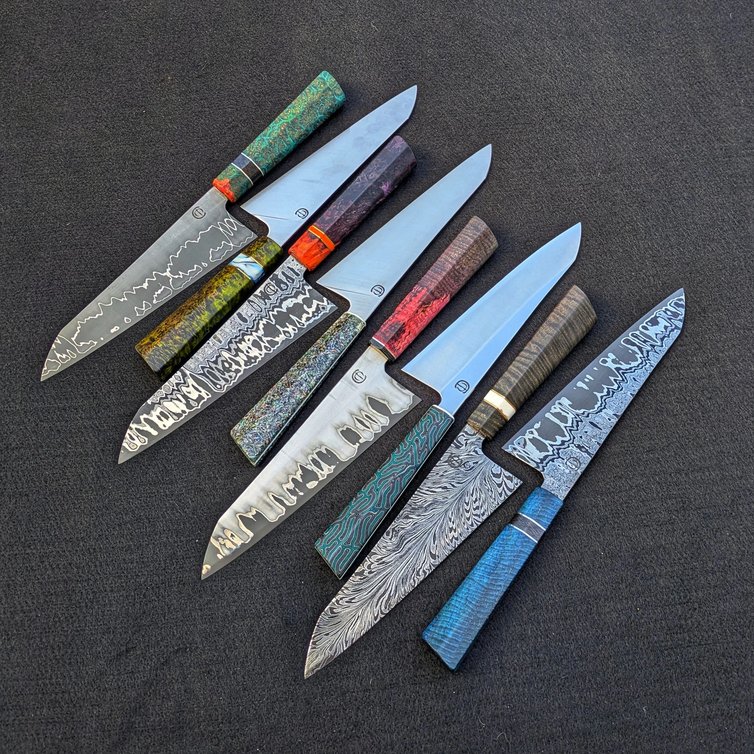 Six decorated kitchen knives with various colorful handles are laid out on a black surface.