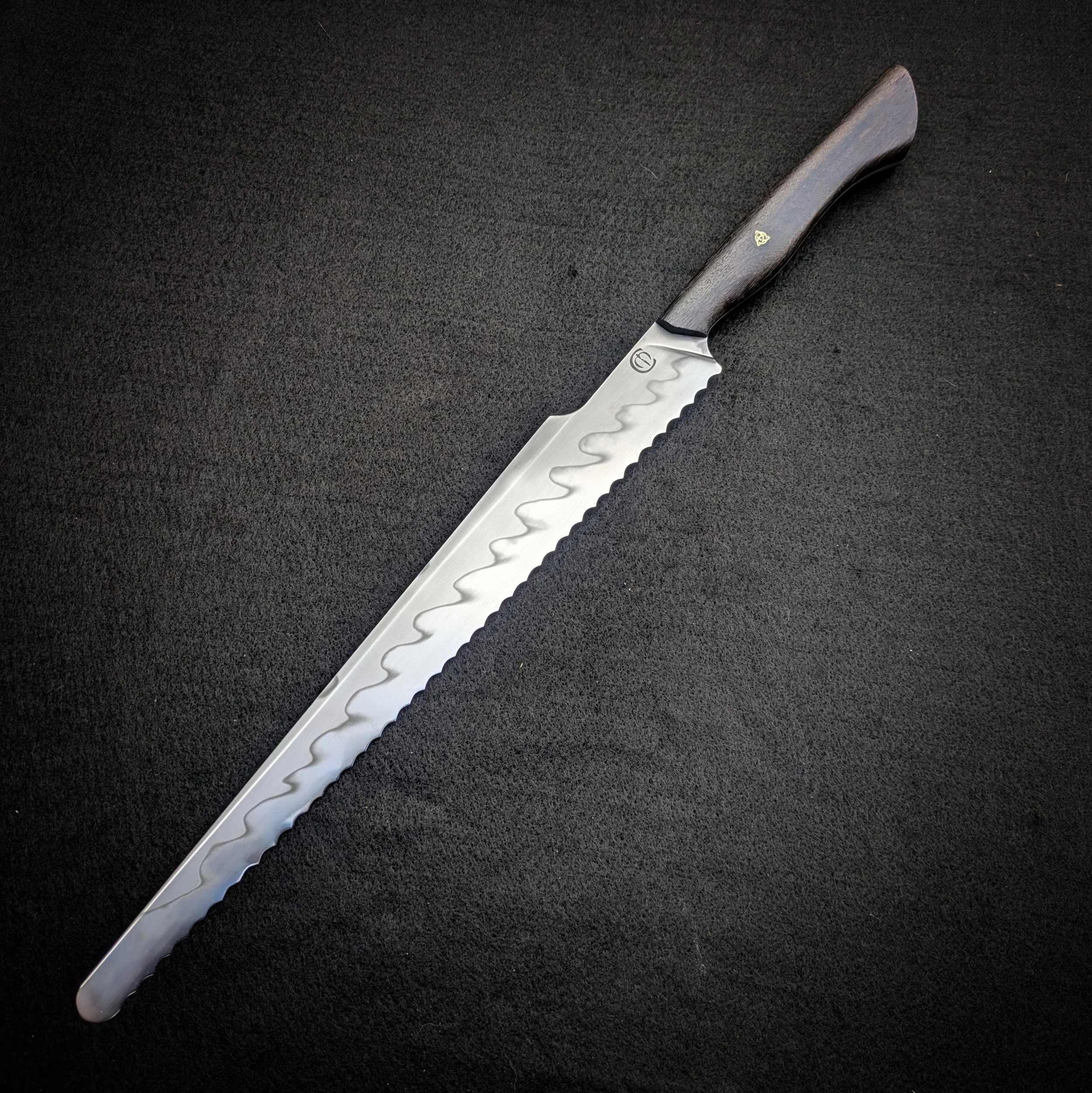 A bread knife with a carbon steel blade and a Bog Oak wooden handle resting on a black textured surface.