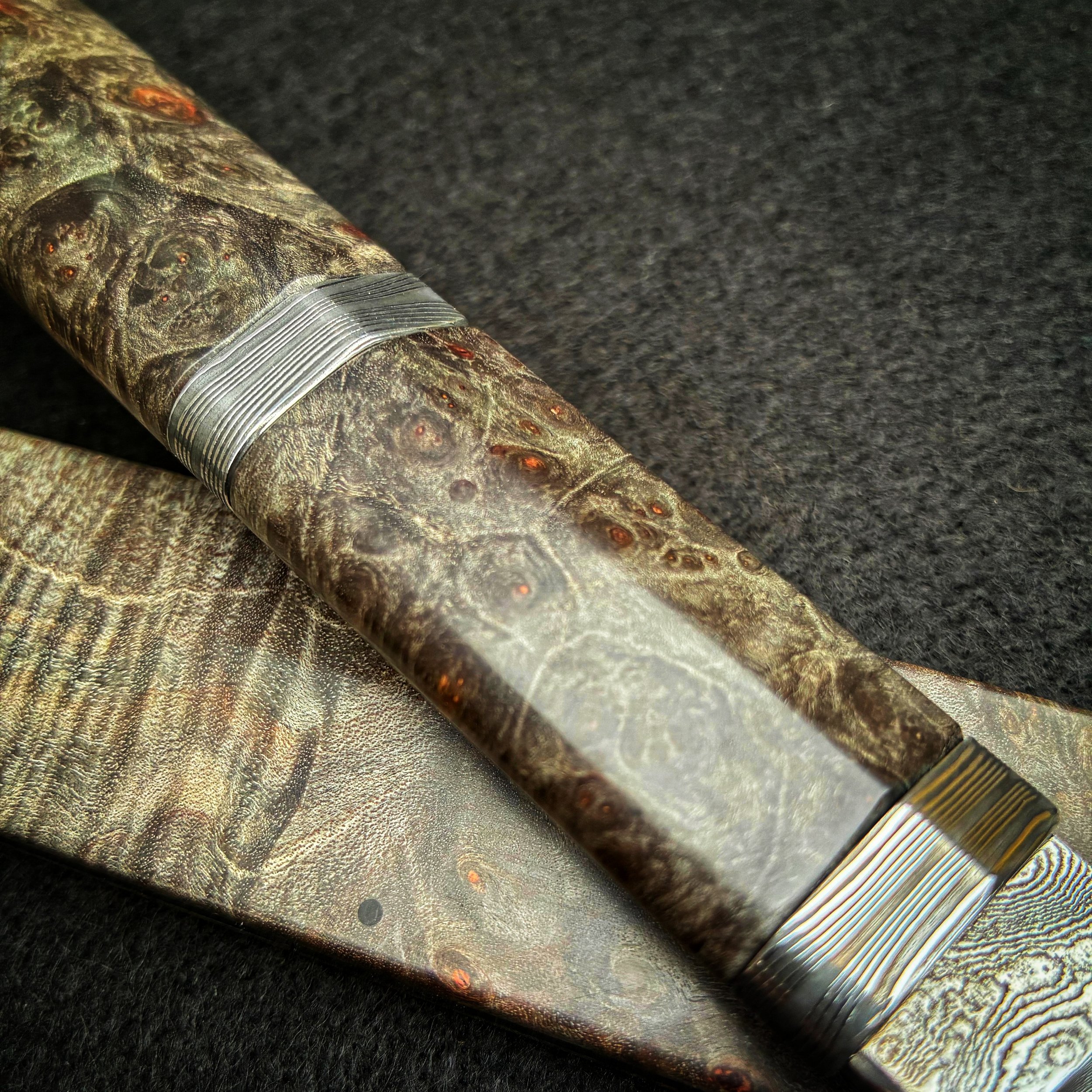 Close-up of a decorative knife with a handle and blade made of patterned material resembling wood grain or camouflage, placed on a dark surface.