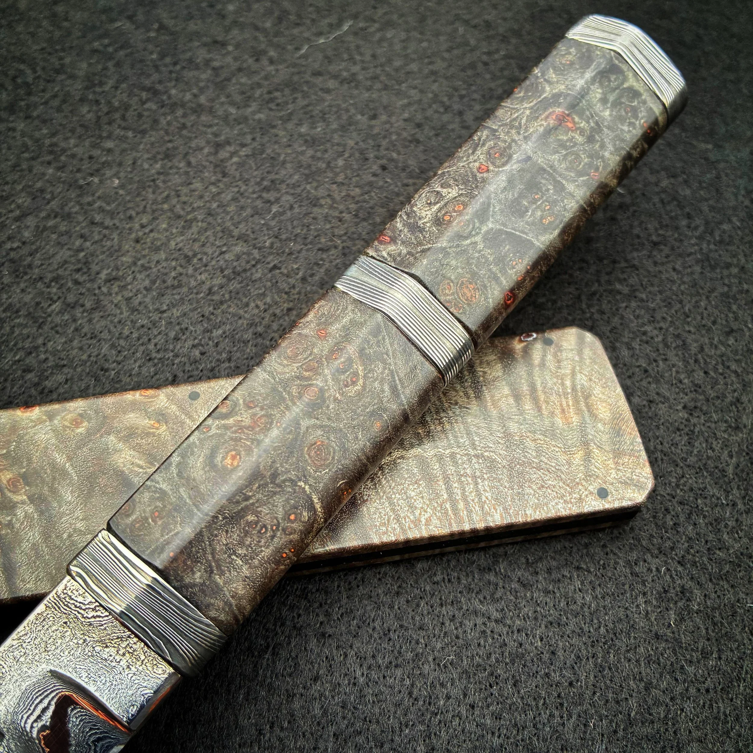 A knife with a handle and blade made from patterned steel, resting on a matching case on a black surface.