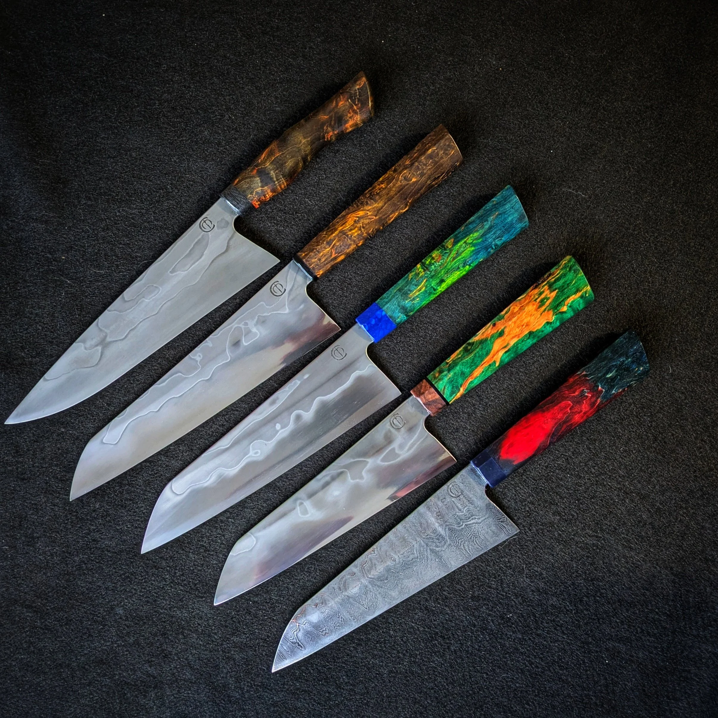Set of five kitchen knives with colorful handles, arranged on a black surface. The handles feature marbled patterns in various colors including orange, green, blue, and red, with a stainless steel blade.
