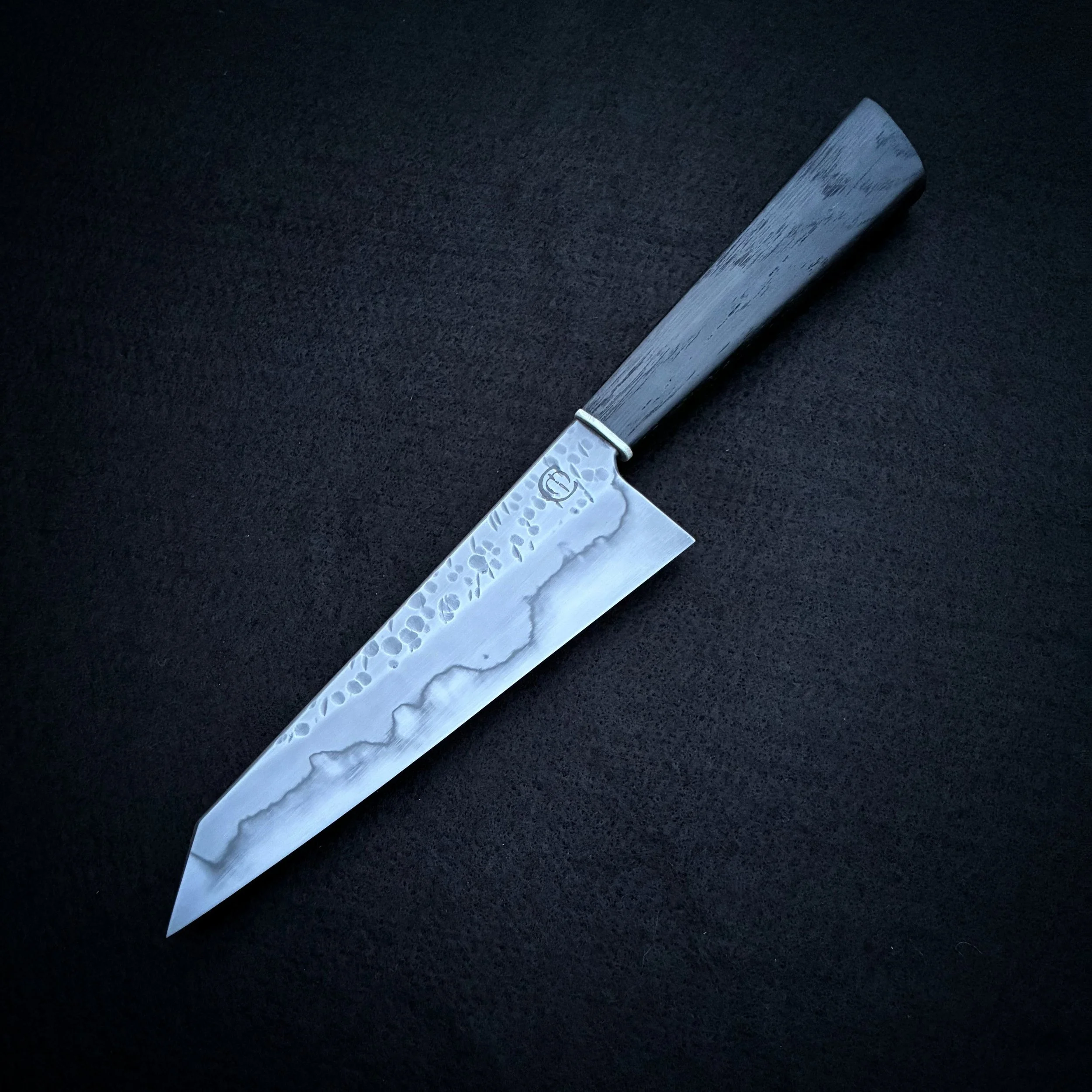 A Japanese-style kitchen knife with a hammered steel blade and a dark wooden handle, positioned diagonally on a black surface.