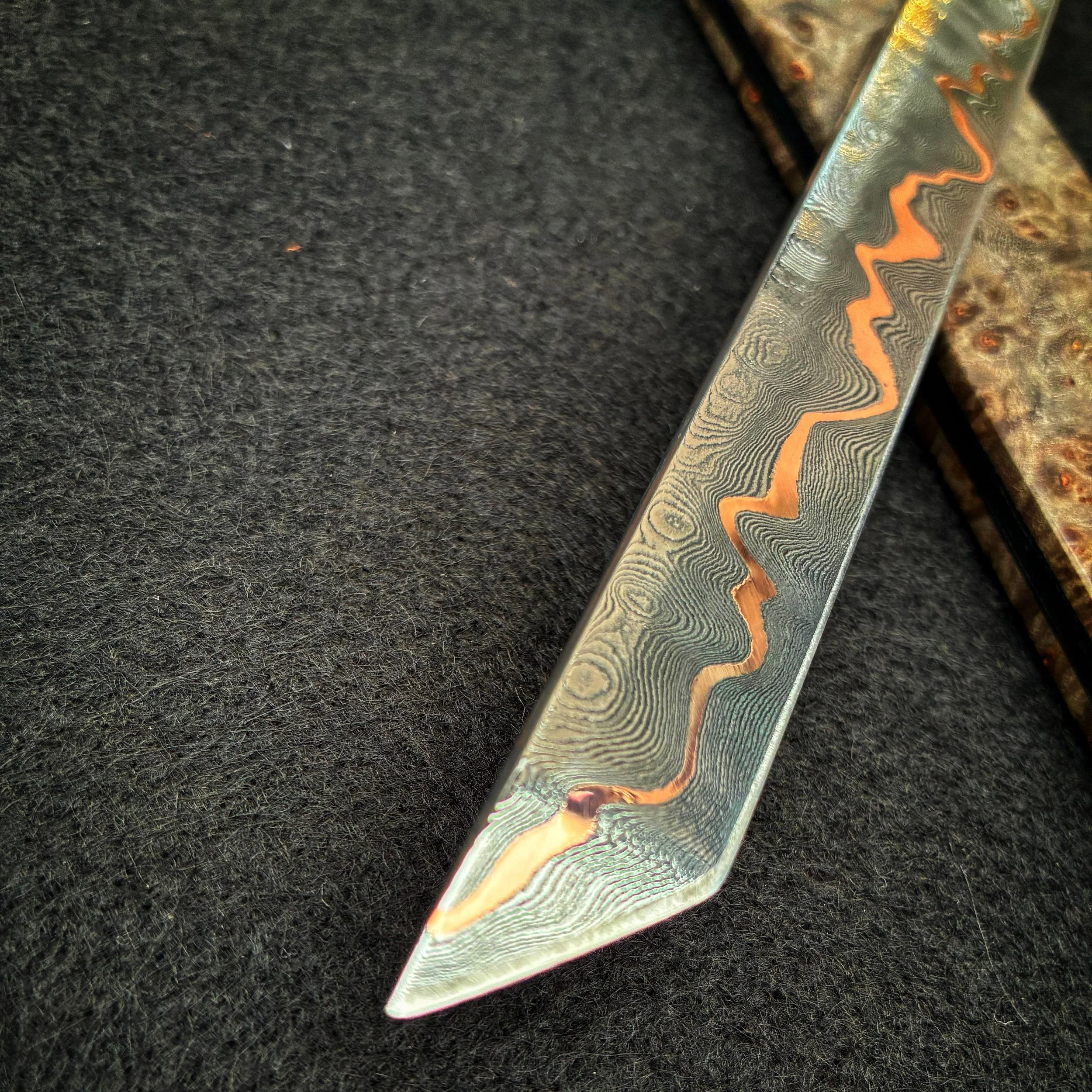Close-up of a decorative metal knife with a detailed, patterned blade showing layers of damascus steel and a copper wavy line.