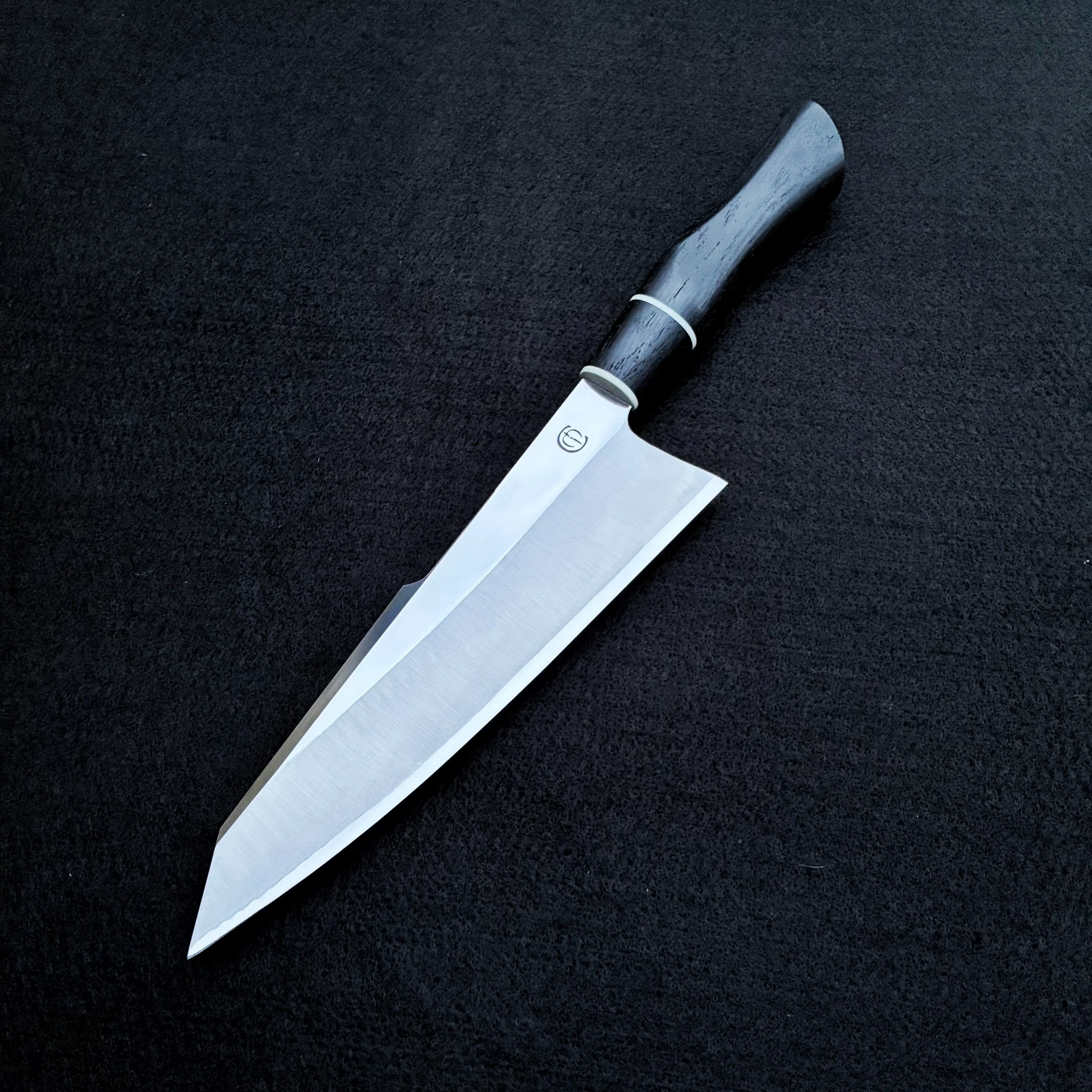 A professional kitchen knife with a dark handle and a broad, sharp blade on a black textured surface.