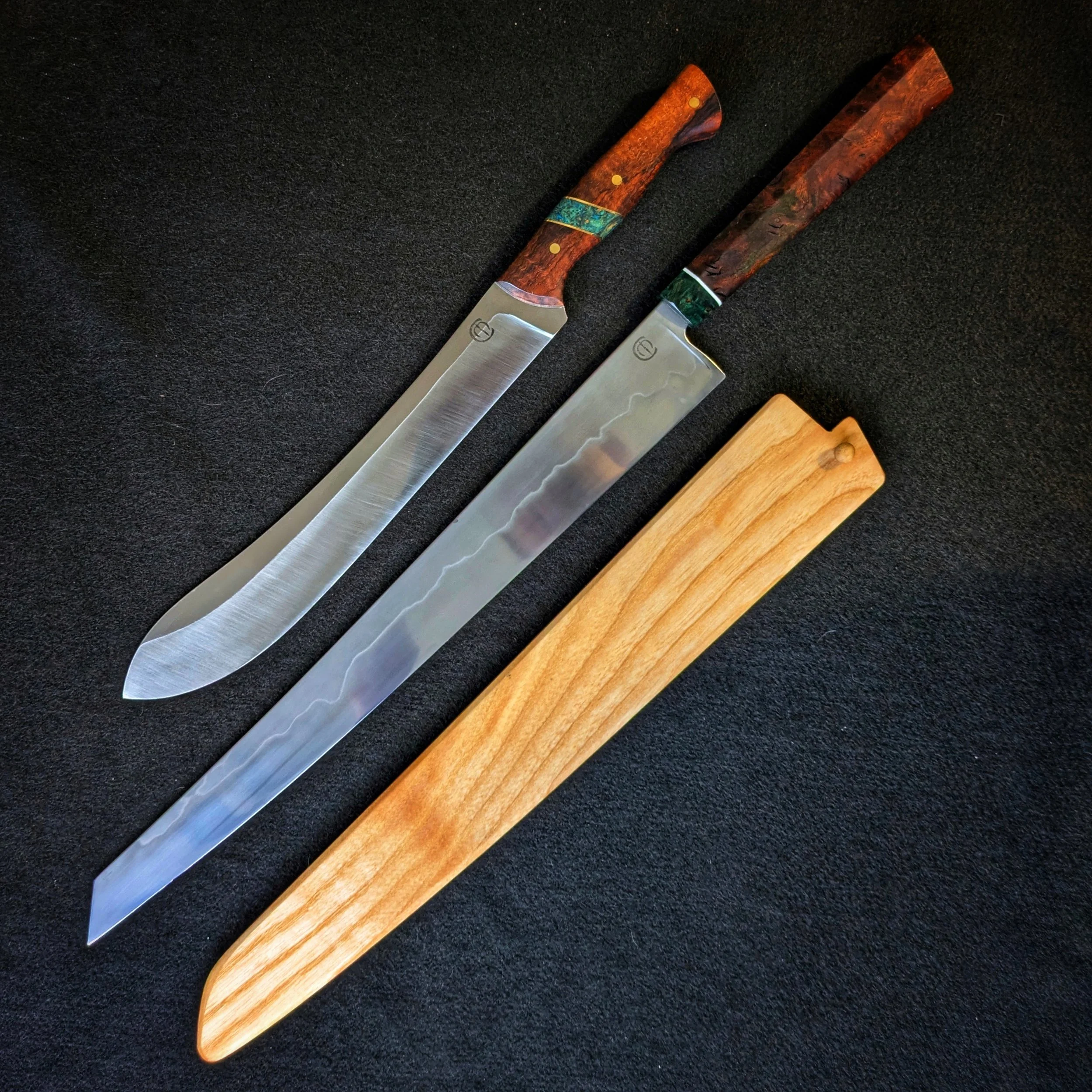 Two knives with wooden handles, one with a silver blade and the other with a pattern on the blade, placed on a black surface with a wooden cutting board partially visible at the bottom.