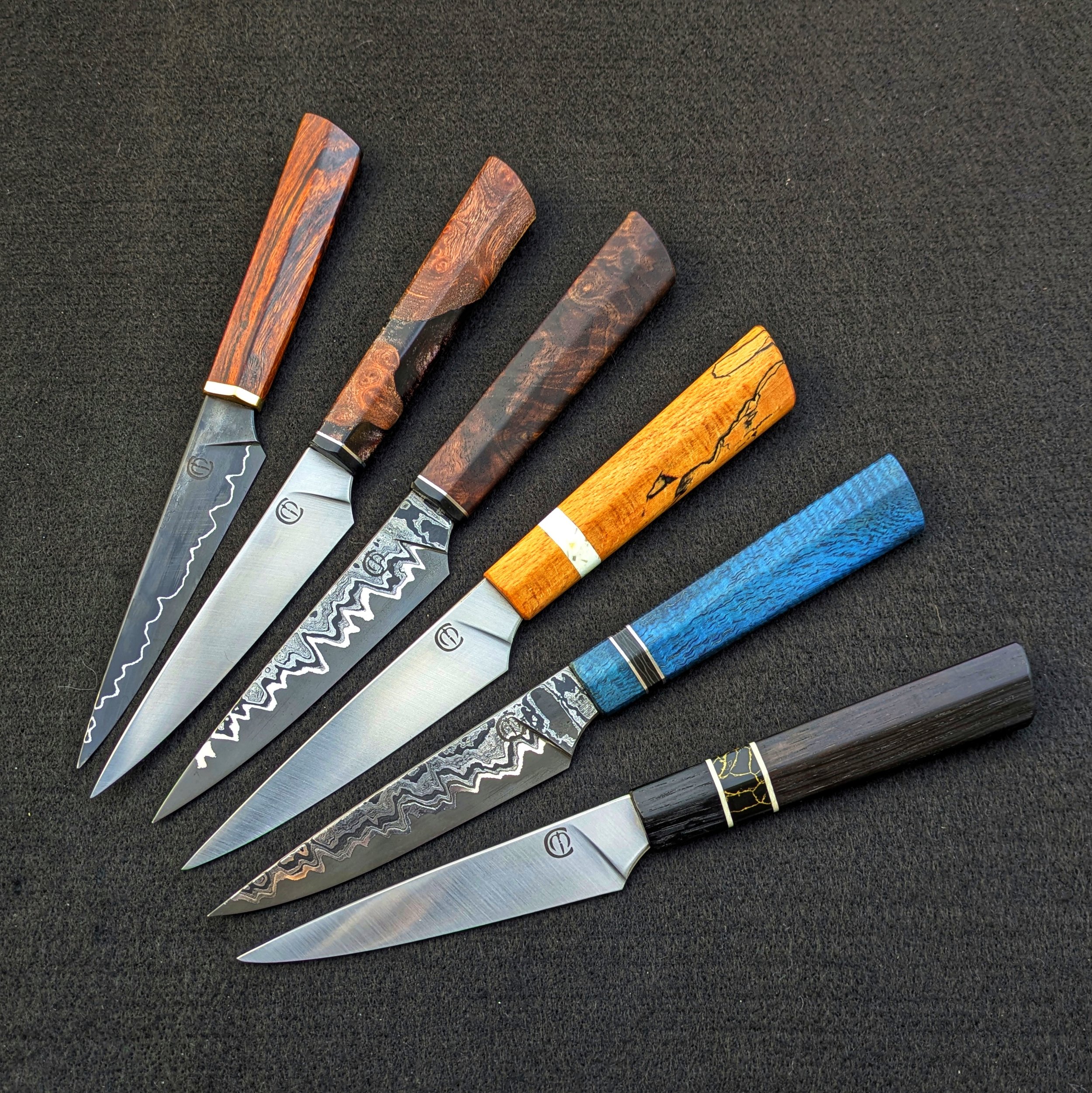 Six handcrafted kitchen knives with decorative wooden handles, arranged on a black surface.