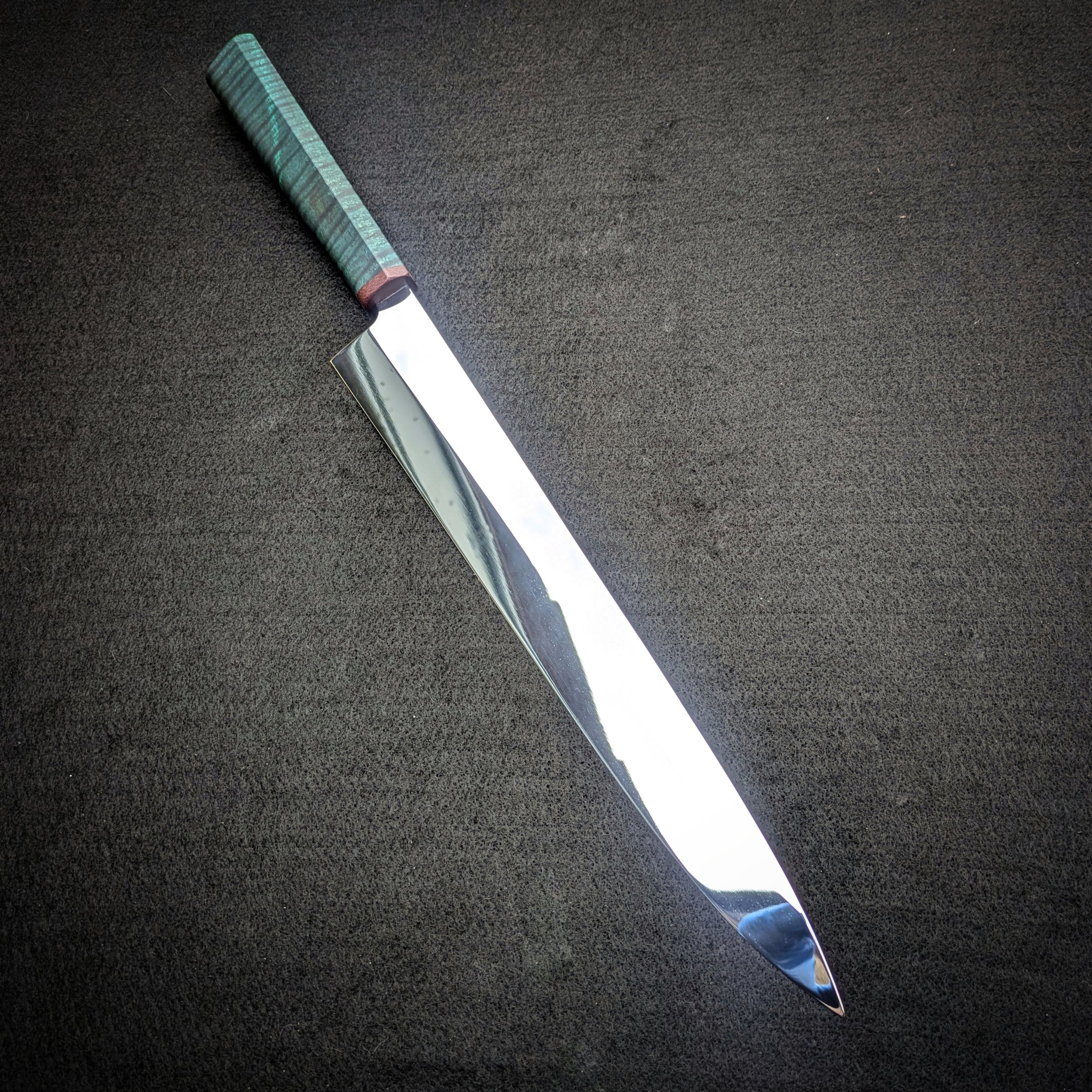 A close-up view of a polished Yanagiba knife with a green handle, resting diagonally on a black textured surface.