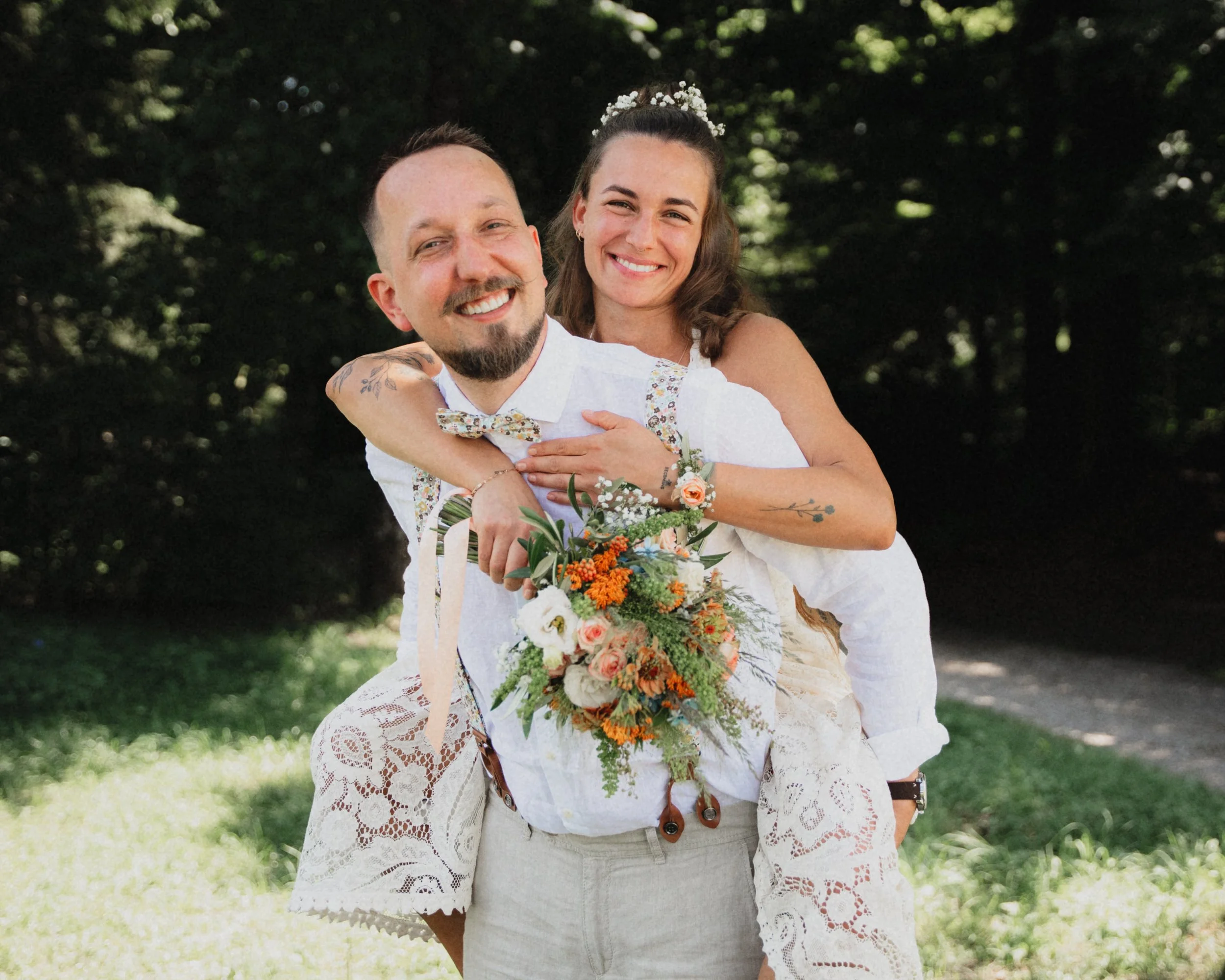 Ein glückliches Paar bei einer Hochzeitsfeier im Freien, das Braut und Bräutigam umarmt. Die Frau trägt ein weißes Kleid mit Blumen und hat einen Blumenkranz im Haar, der Mann trägt ein weißes Hemd mit Fliege mit Blumenmuster.
