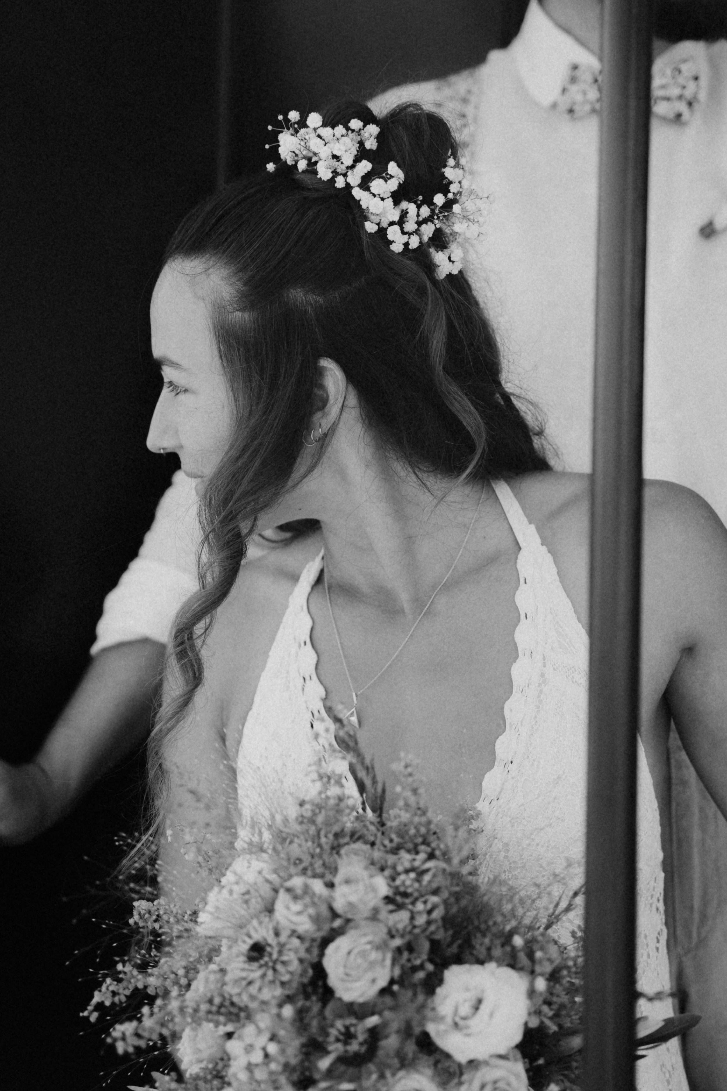 Eine Frau mit dunklen, gewellten Haaren und einem Haarknoten, der mit weißen Blumen verziert ist, trägt ein weißes Spitzenkleid. Sie sitzt neben einem Blumenstrauß mit weißen Blumen, vor einem Spiegel, im Hintergrund ist ein weißer Raum erkennbar.