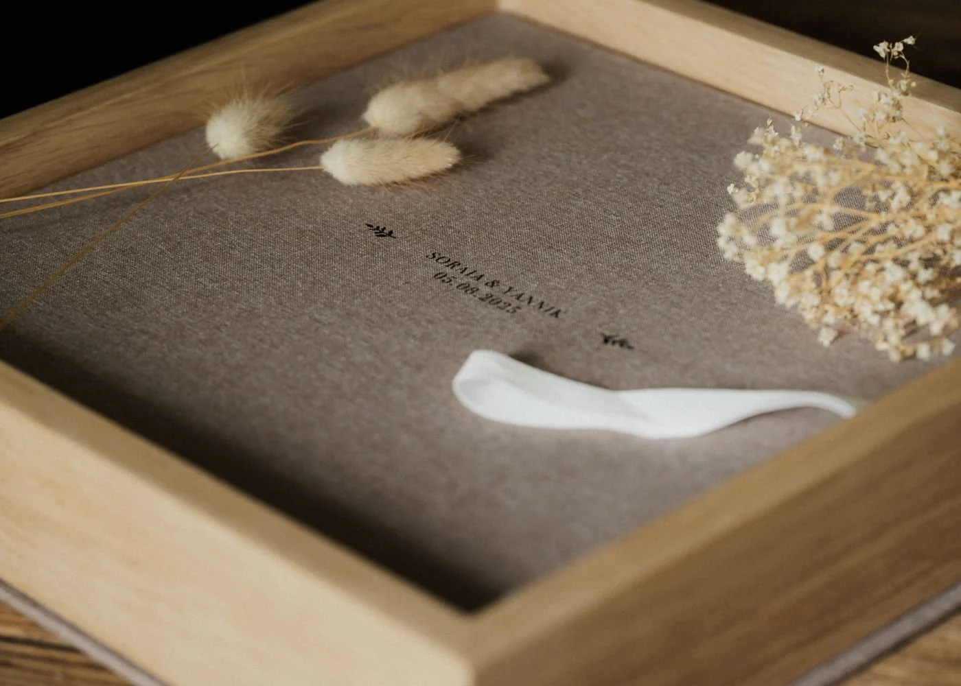 Eine Holz-Box mit einer braunen Innenseite, darin befindet sich eine Information über eine Hochzeit am 02.09.2023, umrahmt von getrockneten Gräsern und Blumen, inklusive einer weißen Schleife.
