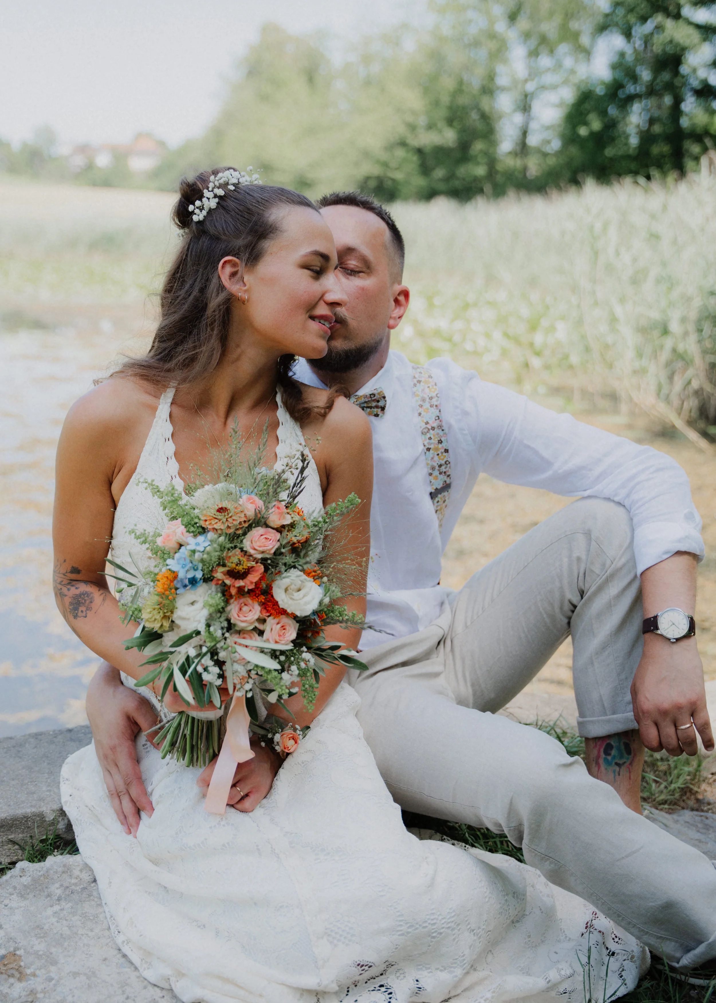 Ein Paar sitzt im Freien, die Frau trägt ein Brautkleid und hält einen Blumenstrauß, der Mann trägt weiße Kleidung und hat bunte Tattoos. Sie scheinen verliebt zu sein.