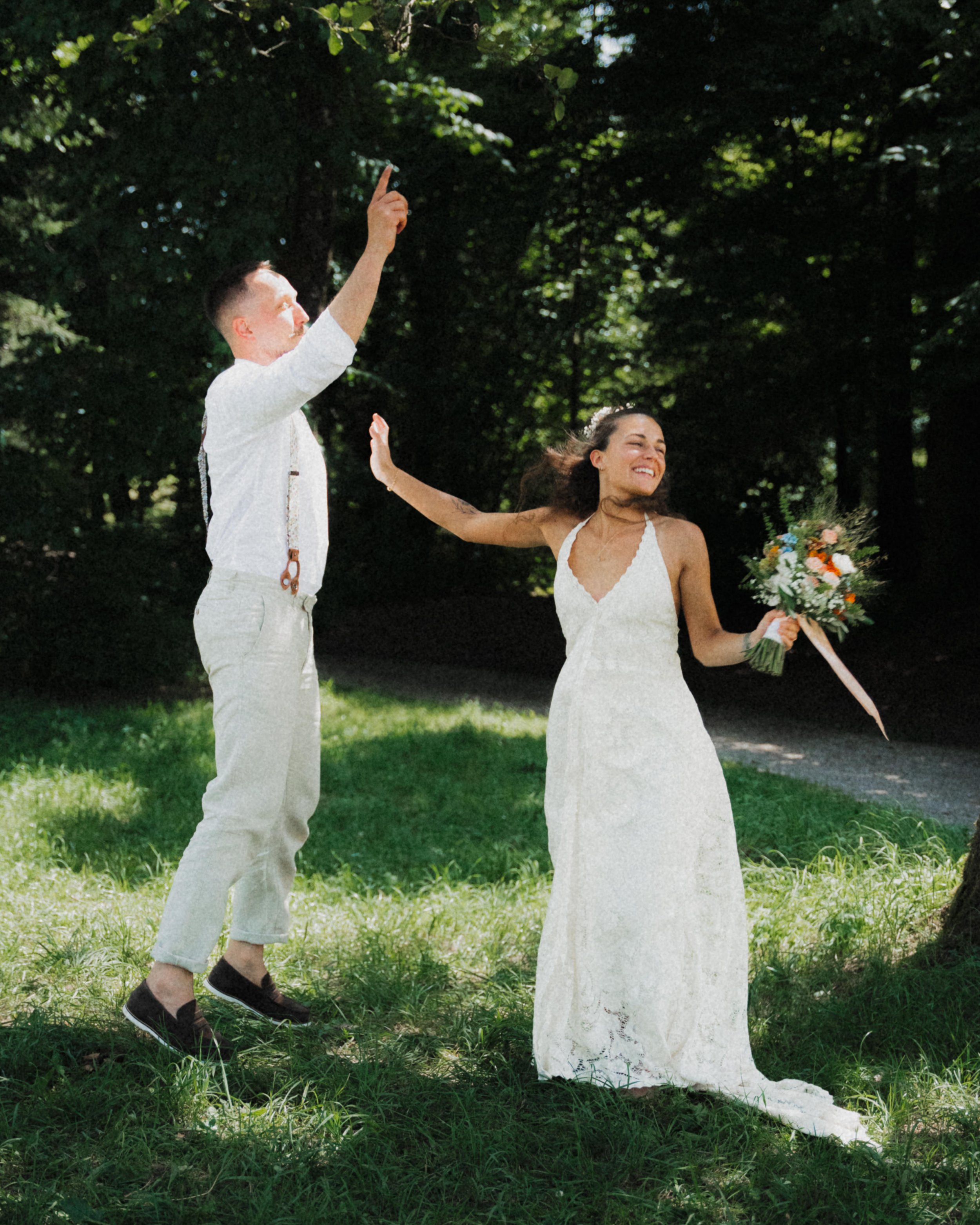 Ein Mann und eine Frau feiern wahrscheinlich eine Hochzeit im Freien. Der Mann trägt helles Hemd, Hosenträger und eine Krawatte, die Frau trägt ein weißes Brautkleid und hält einen Blumenstrauß. Beide lächeln und freuen sich, während sie in die Luft 