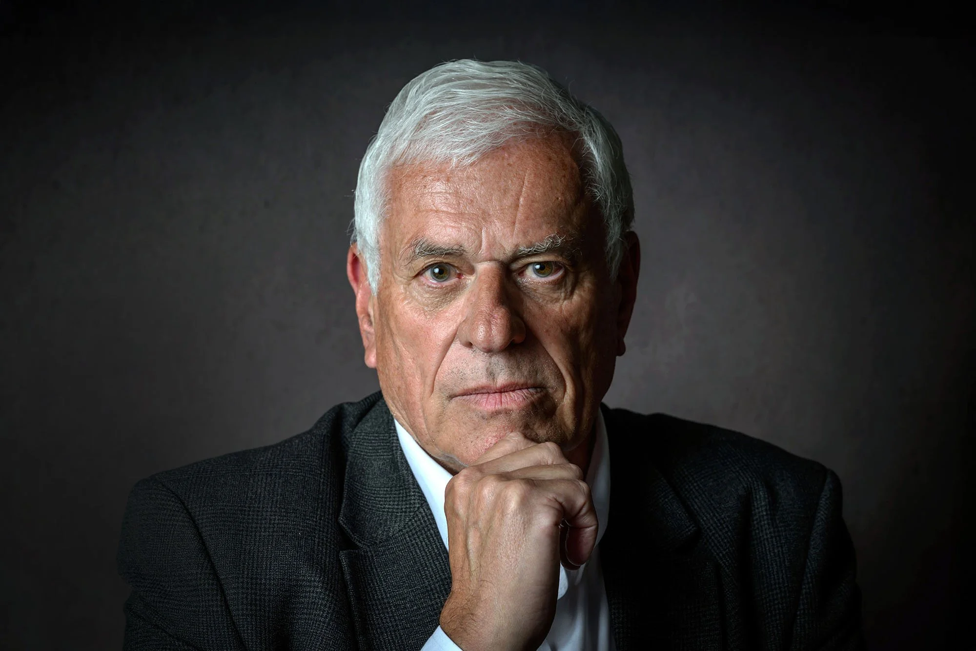 Close-up portrait of an elderly man with white hair, wearing a black suit and white shirt, against a dark background. He is looking directly at the camera with a contemplative expression, resting his chin on his hand.