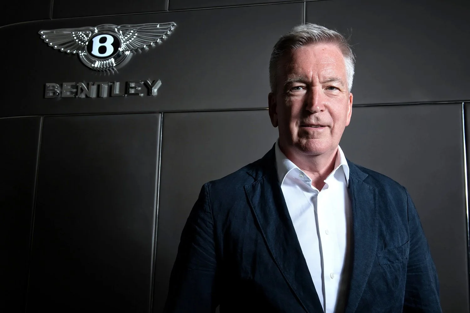 A man in a white shirt and navy blazer standing in front of a black wall with the Bentley logo and a decorative winged emblem.
