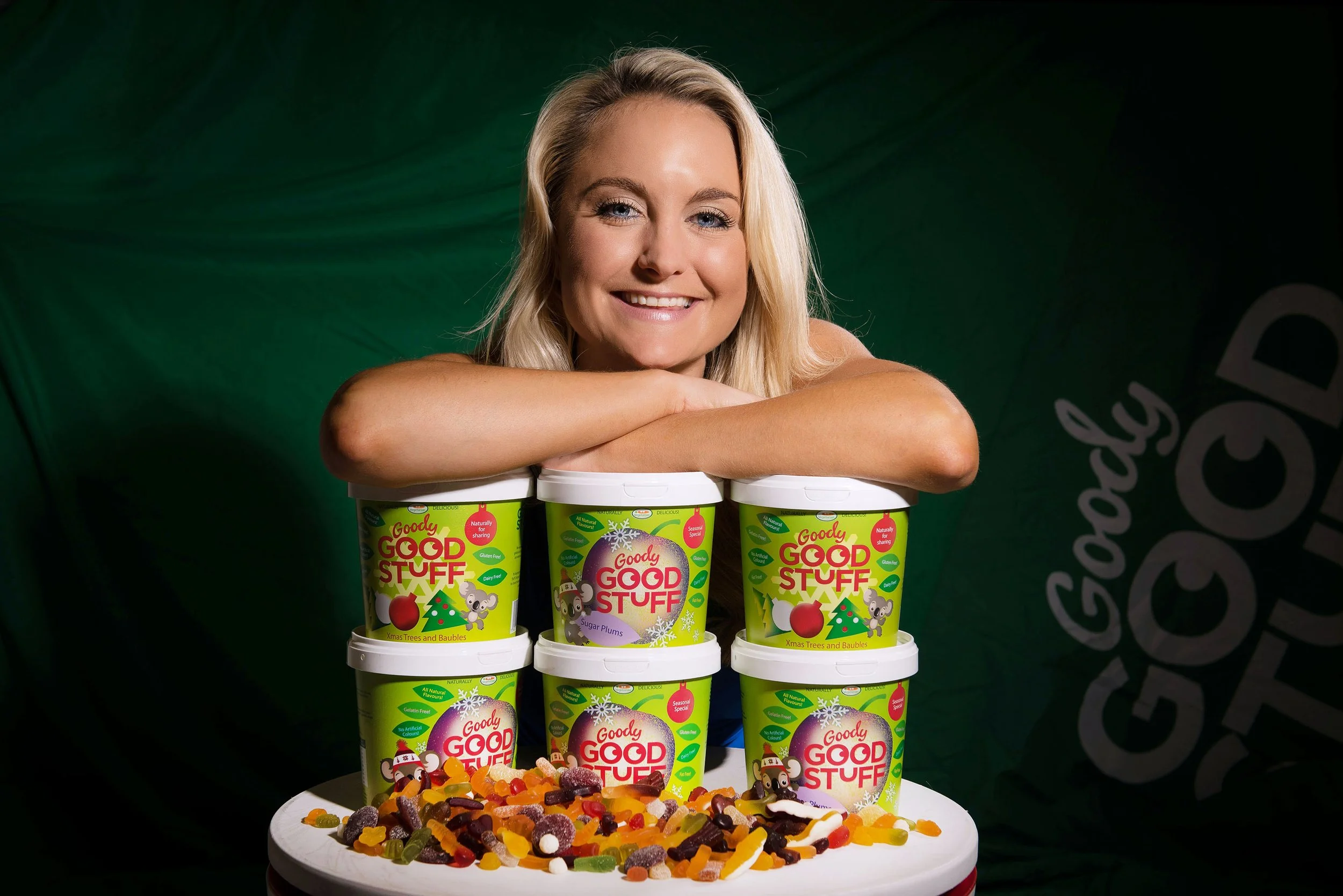 A woman smiles resting her chin on folded arms above six cartons of Goody Good Stuff candy, displayed on a white cake stand, with gummy candies spread around.
