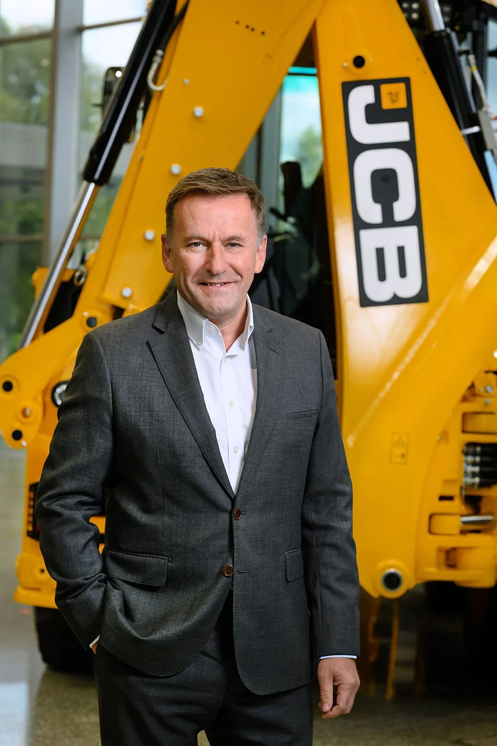 A man dressed in a gray suit with a white shirt stands indoors in front of a large yellow JCB construction vehicle.