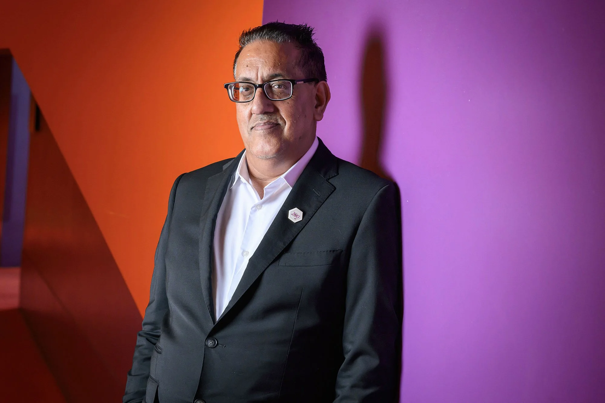 Portrait of a man in a suit and glasses standing against a vibrant, colorful background involving purple and orange hues.