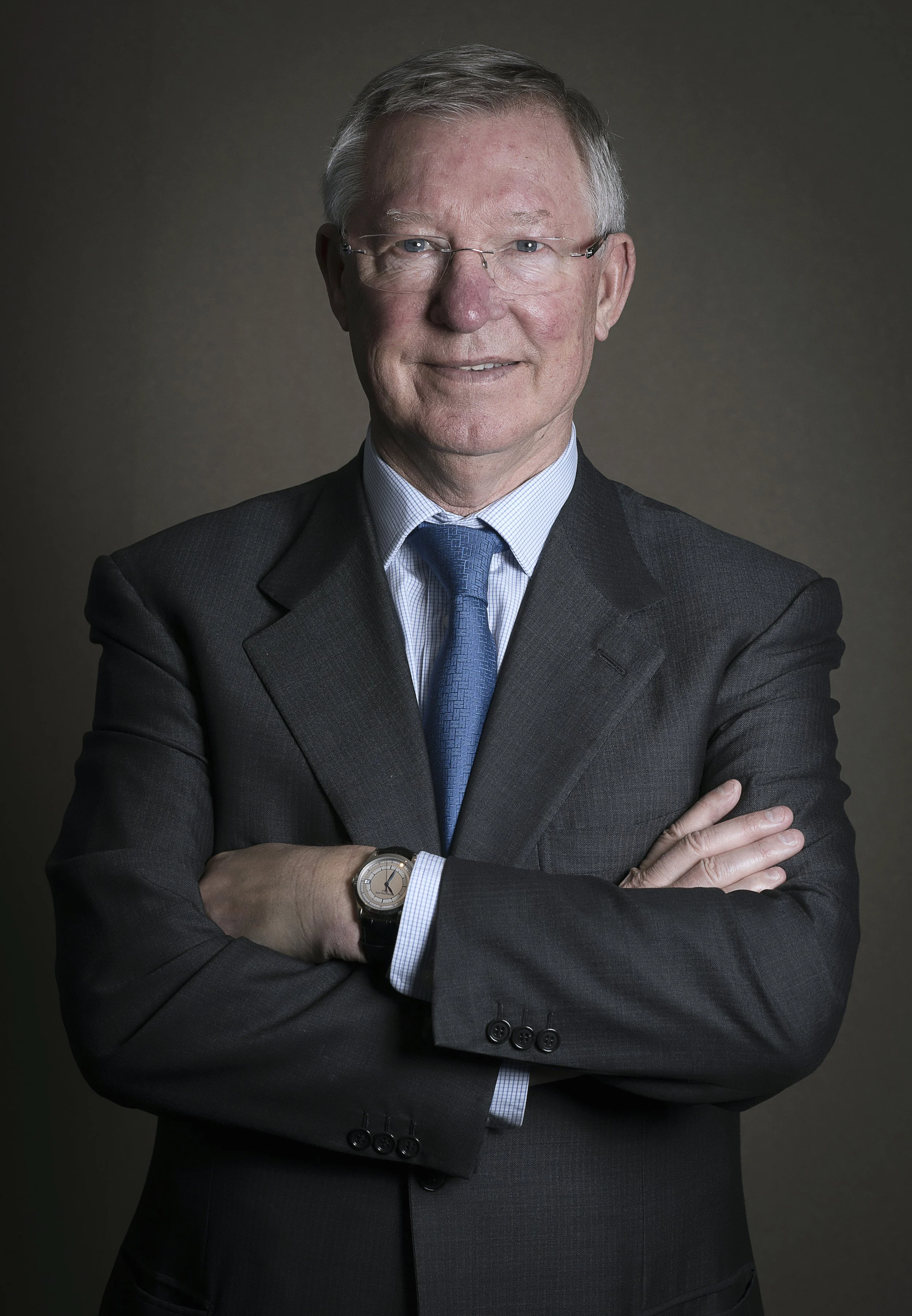 Portrait of an older man in a suit with glasses, crossing his arms, against a dark background.