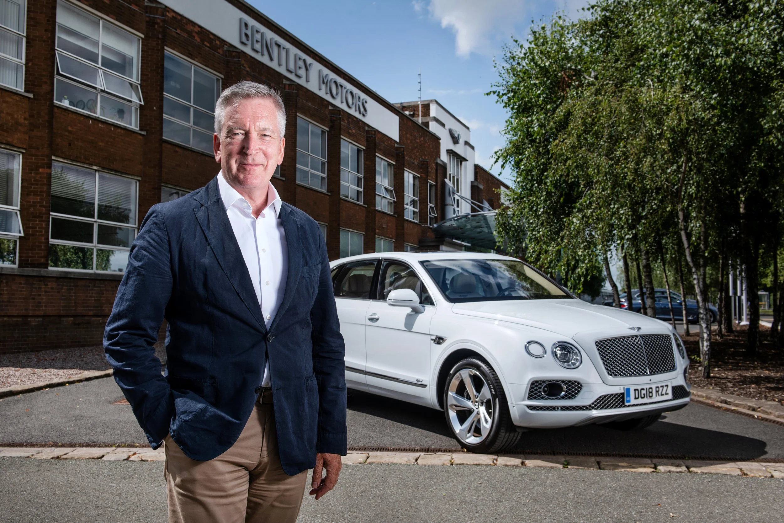 A man in a navy blazer and beige pants standing in front of a white luxury car parked in front of a brick building with large windows and a sign that reads 'BENTLEY MOTORS'. There are trees and blue sky in the background.
